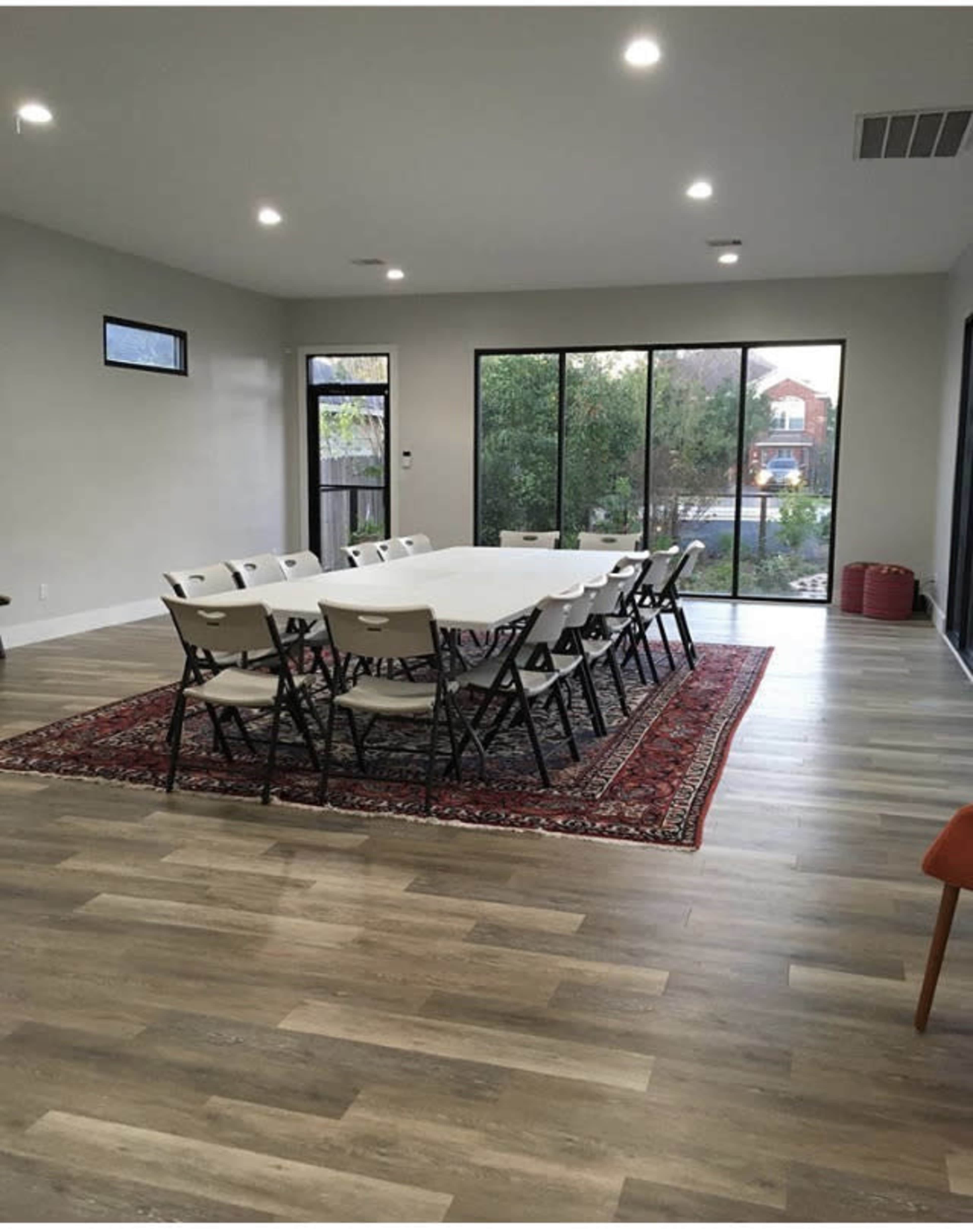 A spacious room features a long table surrounded by folding chairs, with large windows offering a view of greenery outside.