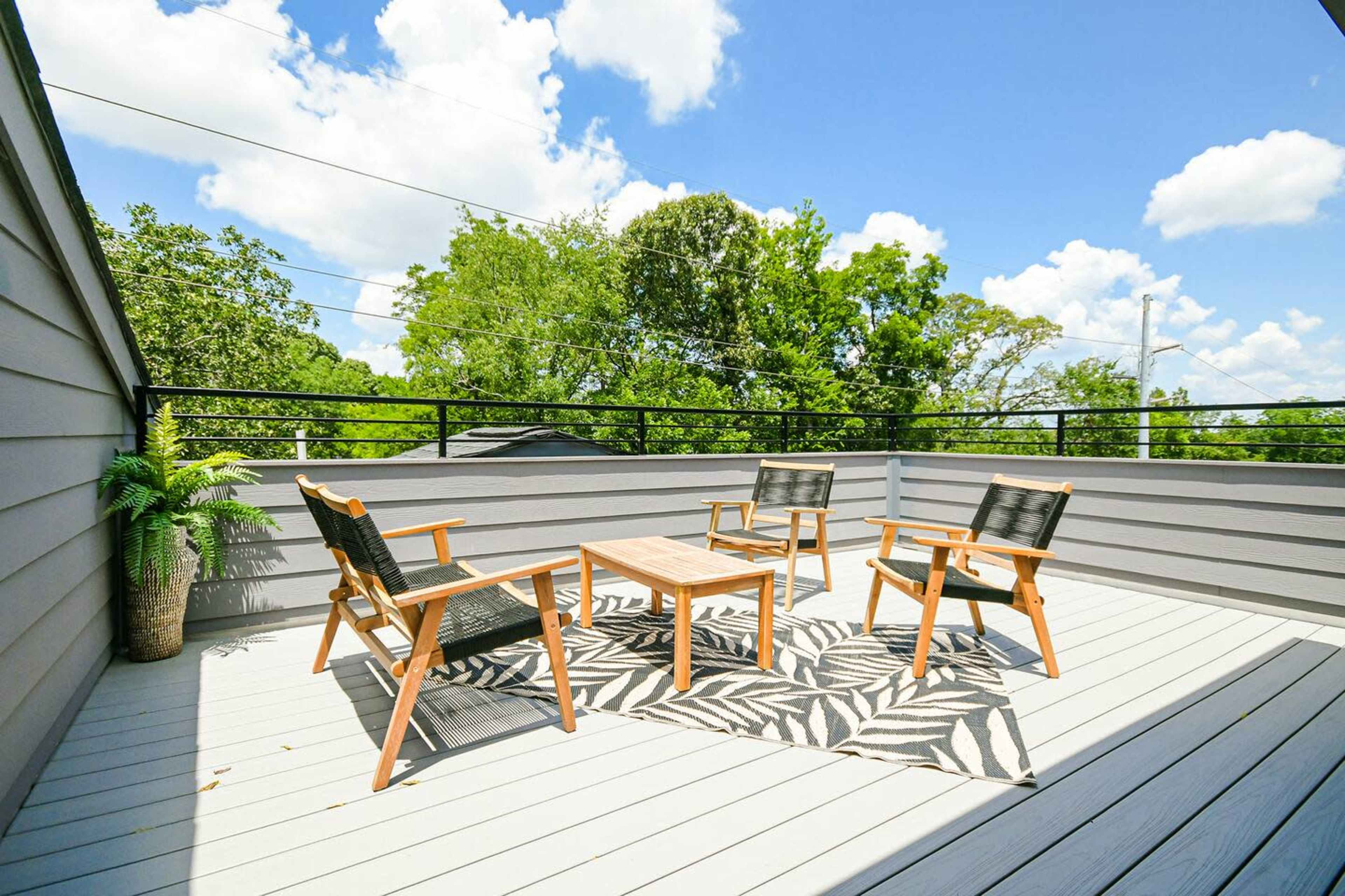 A rooftop terrace features four wooden chairs and a table arranged on a patterned rug, surrounded by greenery and a clear blue sky.