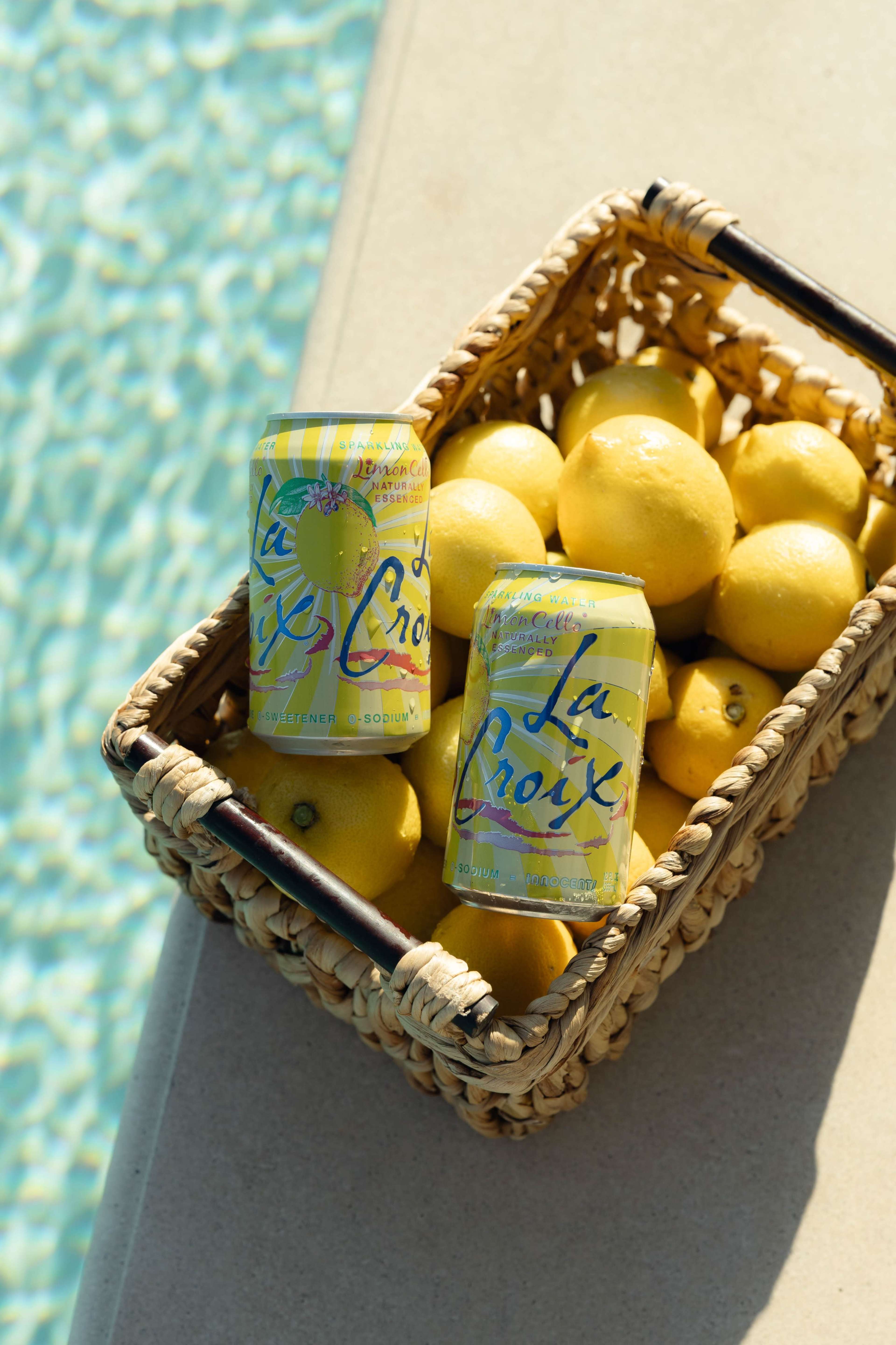 A wicker basket filled with lemons and two cans of La Croix sits beside a pool.