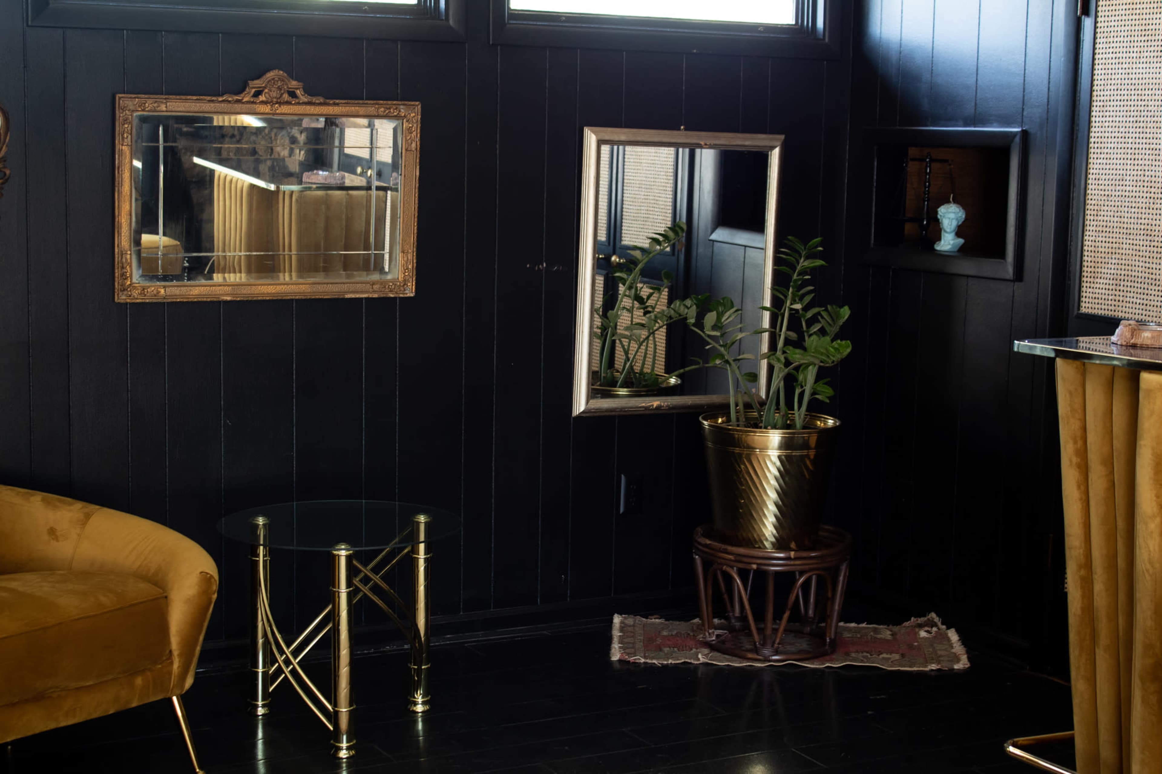 The scene features a dark-walled interior with a gold-accented armchair, a mirrored wall, and a potted plant on a wooden stand.
