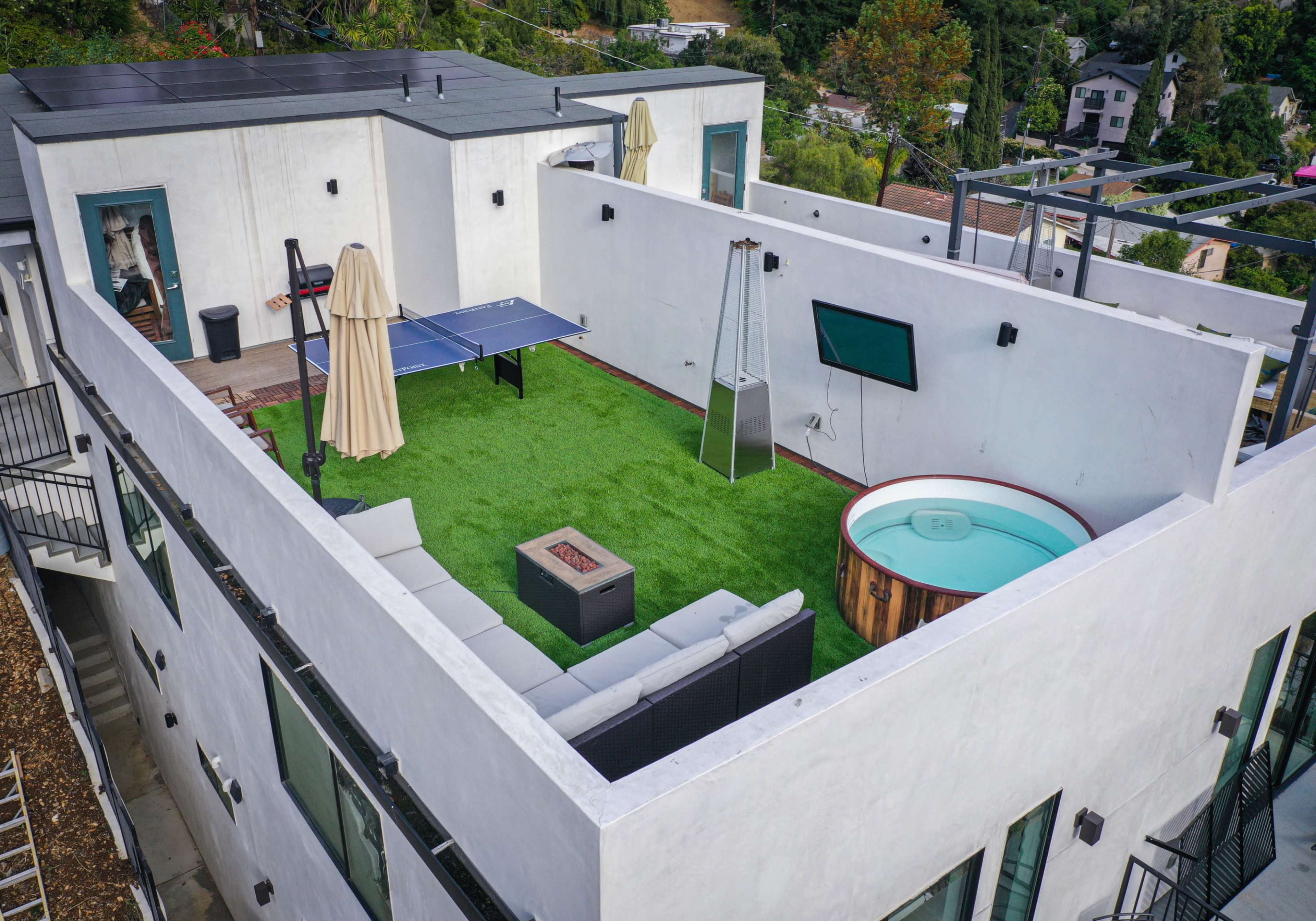 A rooftop terrace featuring a green artificial lawn, a ping pong table, a small hot tub, a television mounted on a wall, and a seating area with modern furniture.