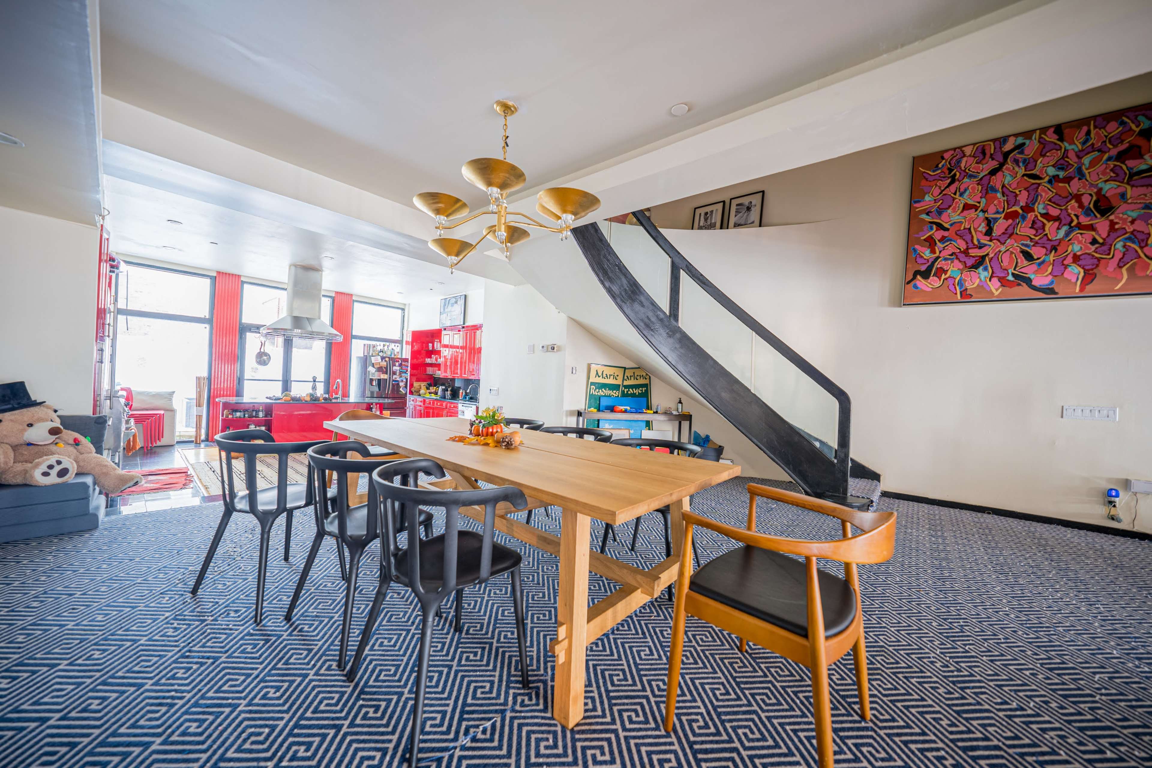 A modern dining area with a wooden table surrounded by black and wooden chairs, a staircase in the background, and colorful decor on the walls.