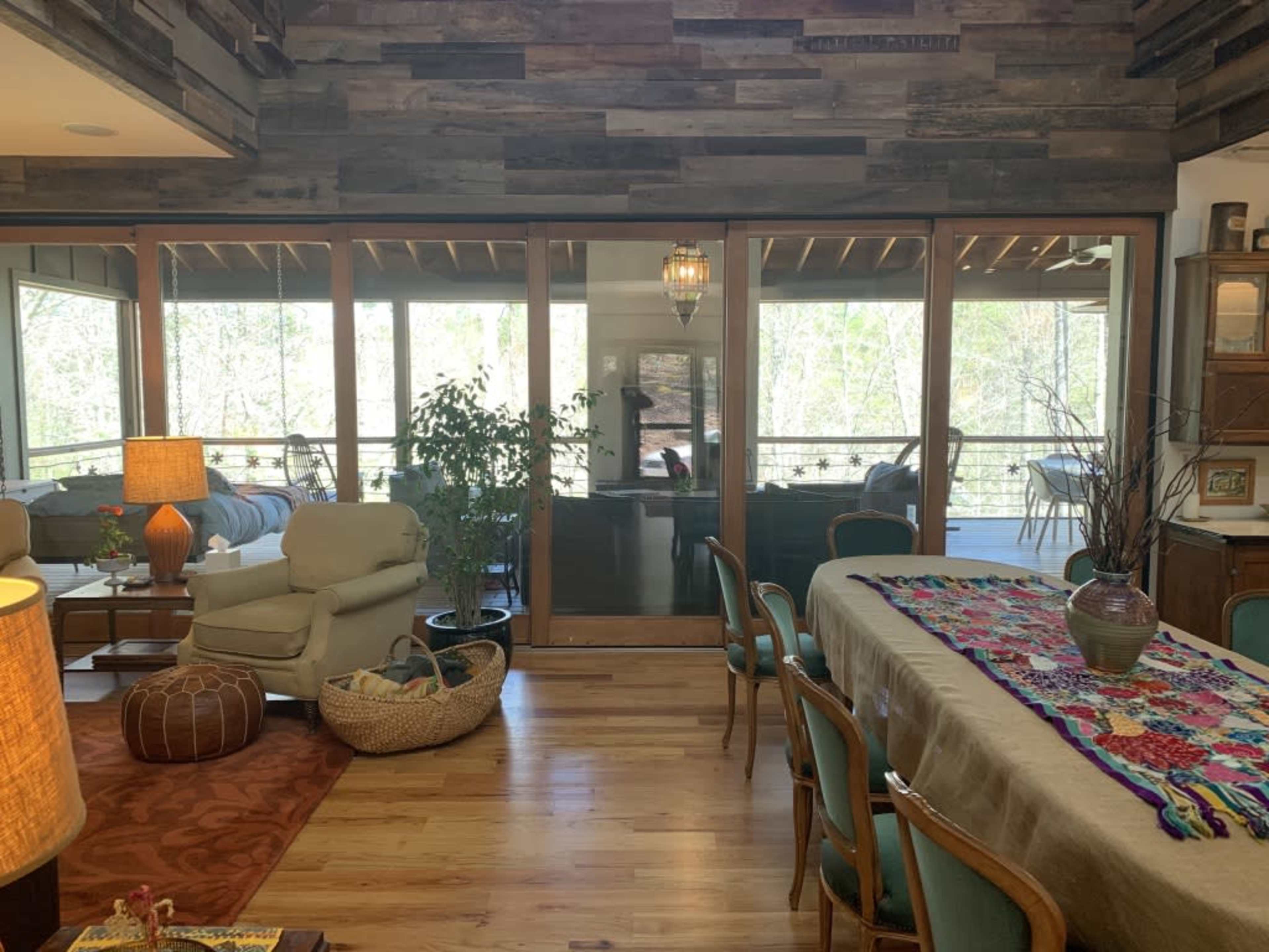 A spacious living area with wooden walls, large windows, and a dining table adorned with a colorful tablecloth overlooks a balcony surrounded by greenery.