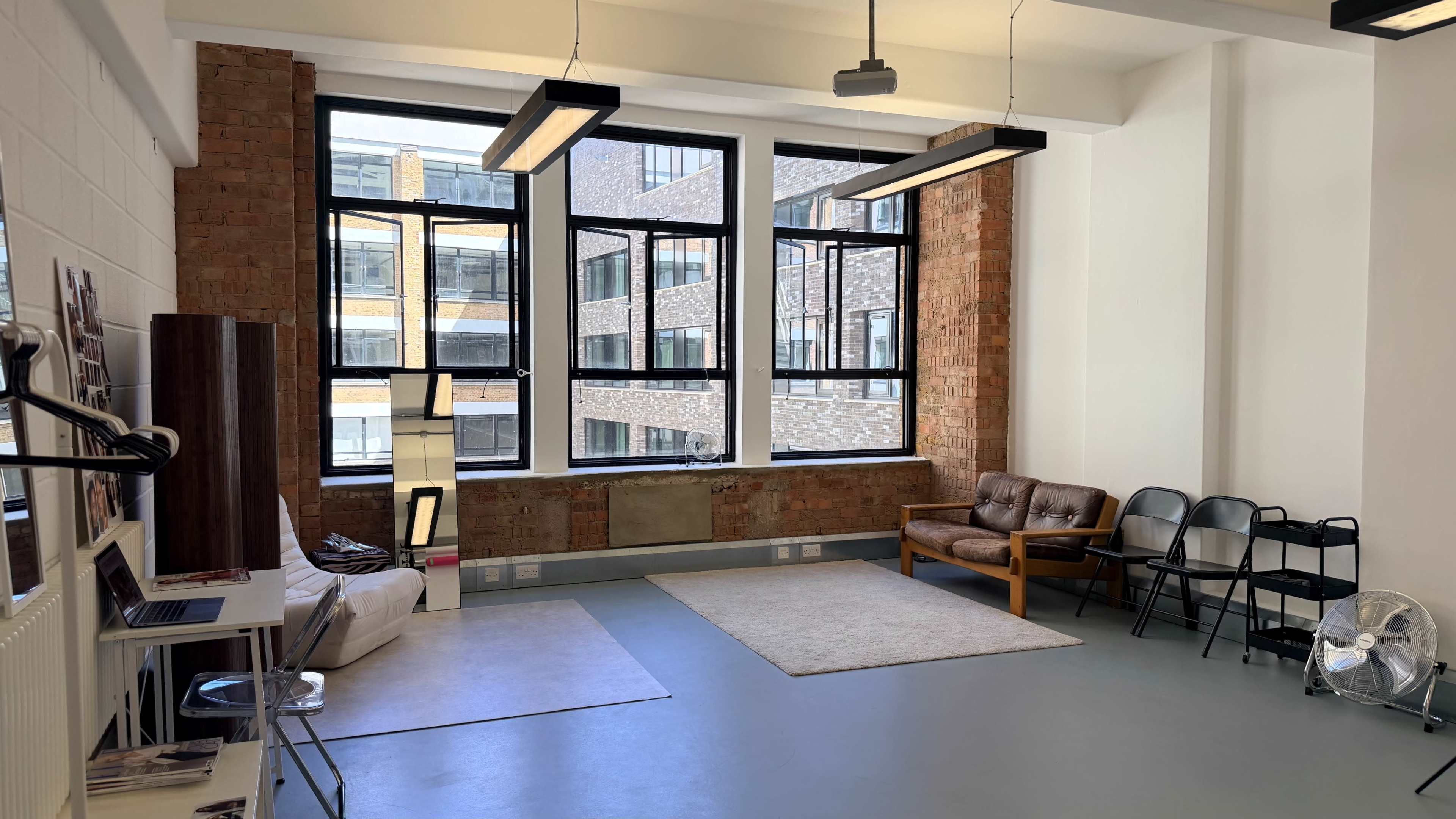The image shows a spacious room with large windows, exposed brick walls, and a combination of seating options, including a brown leather sofa and black chairs.