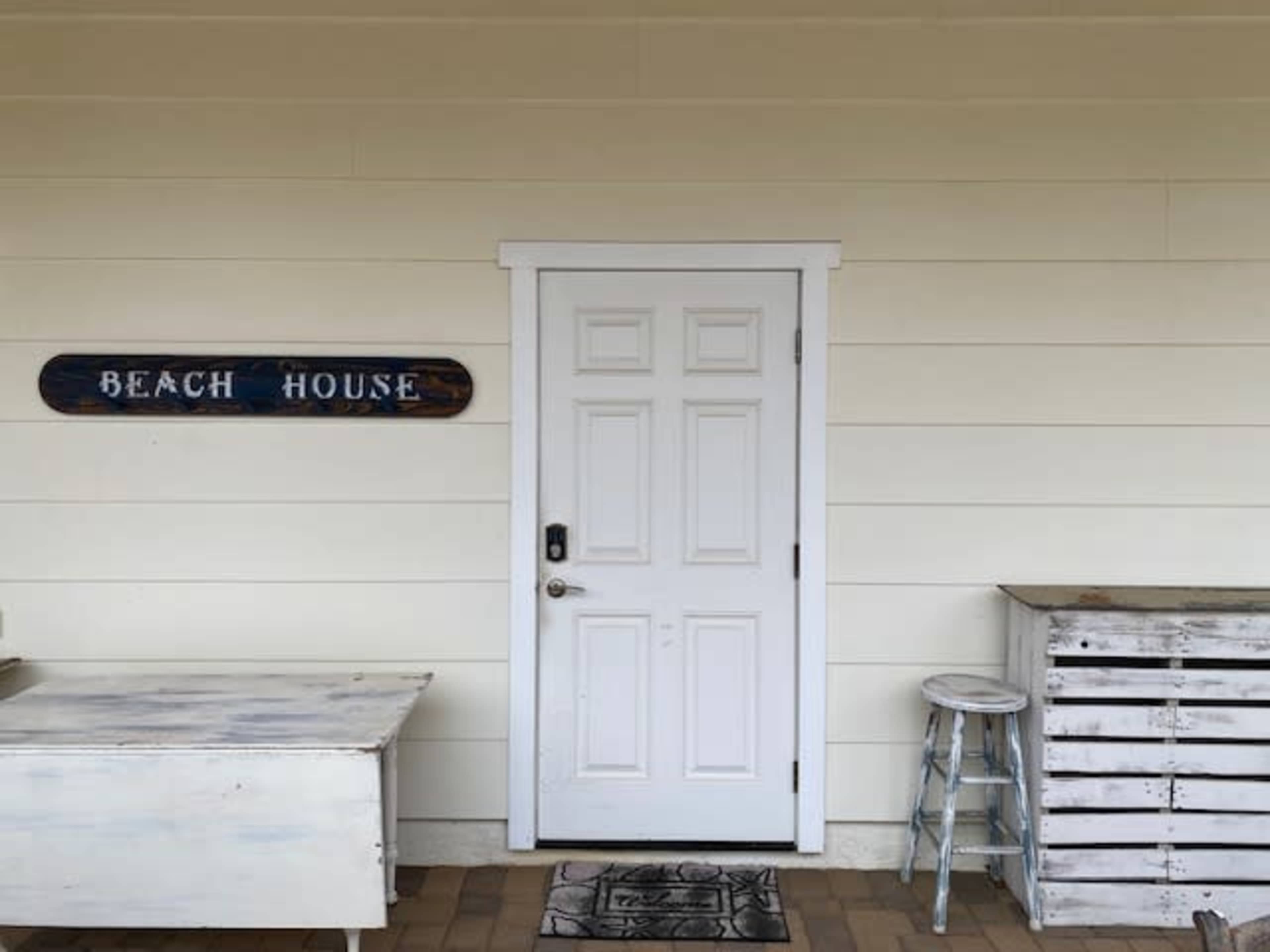 A white door with a black handle is set into a light-colored wooden wall, accompanied by a "BEACH HOUSE" sign and two rustic wooden furniture pieces.