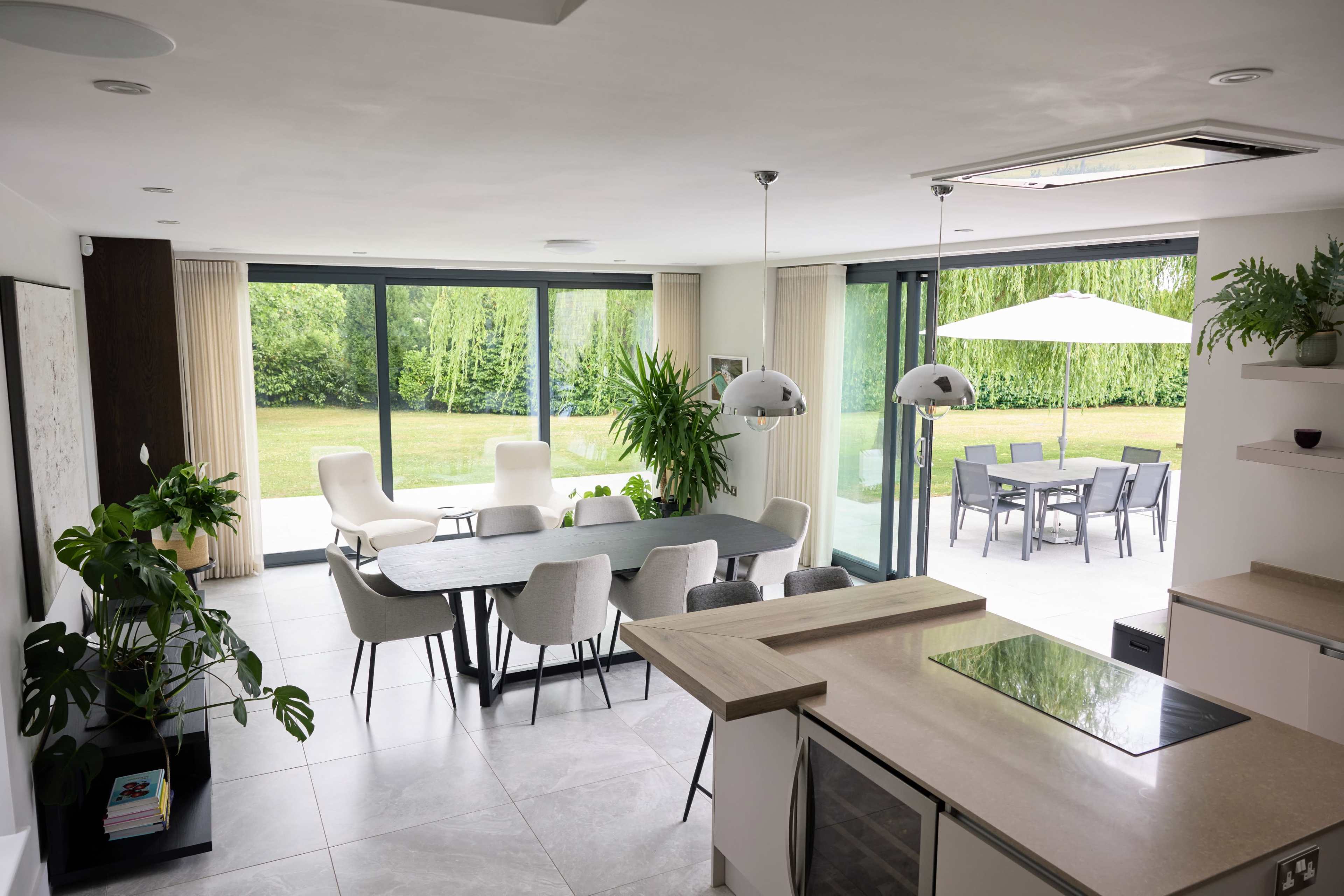 A modern dining area with a large table and chairs overlooks a spacious patio surrounded by greenery.