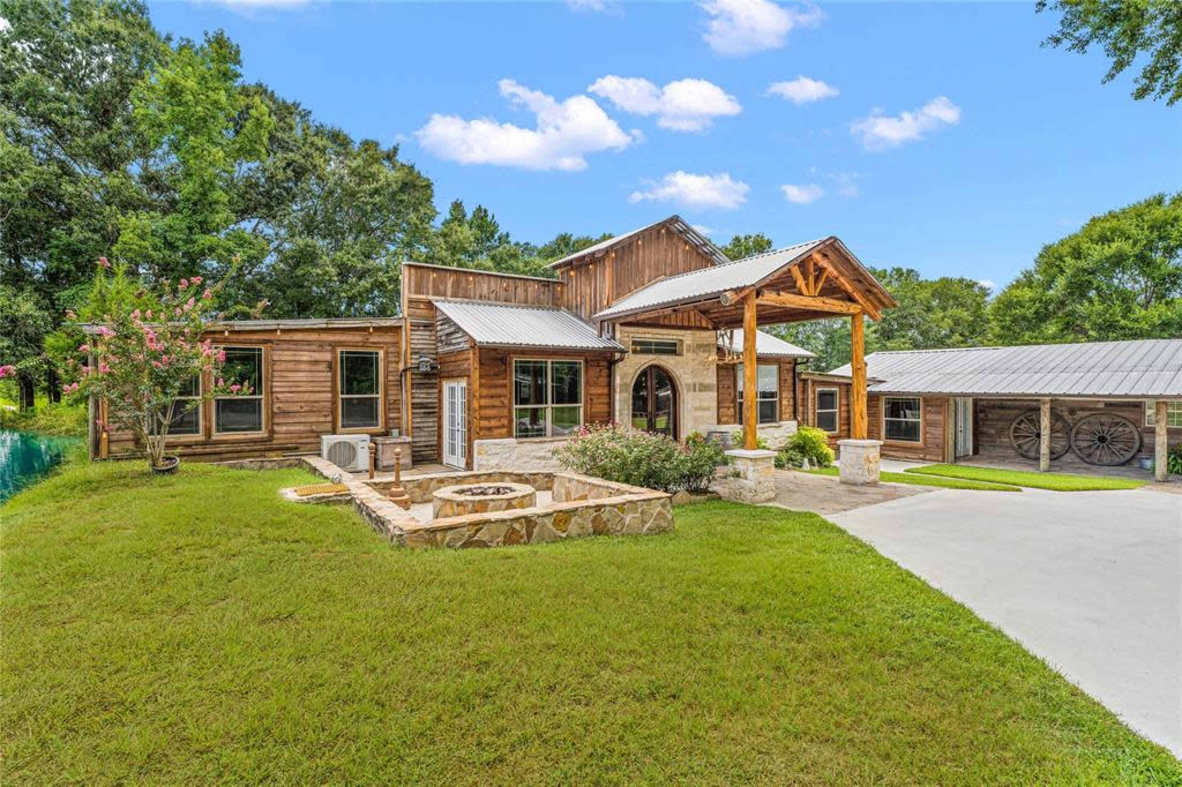 A rustic-style wooden house with a large front porch, surrounded by greenery and a gravel driveway.