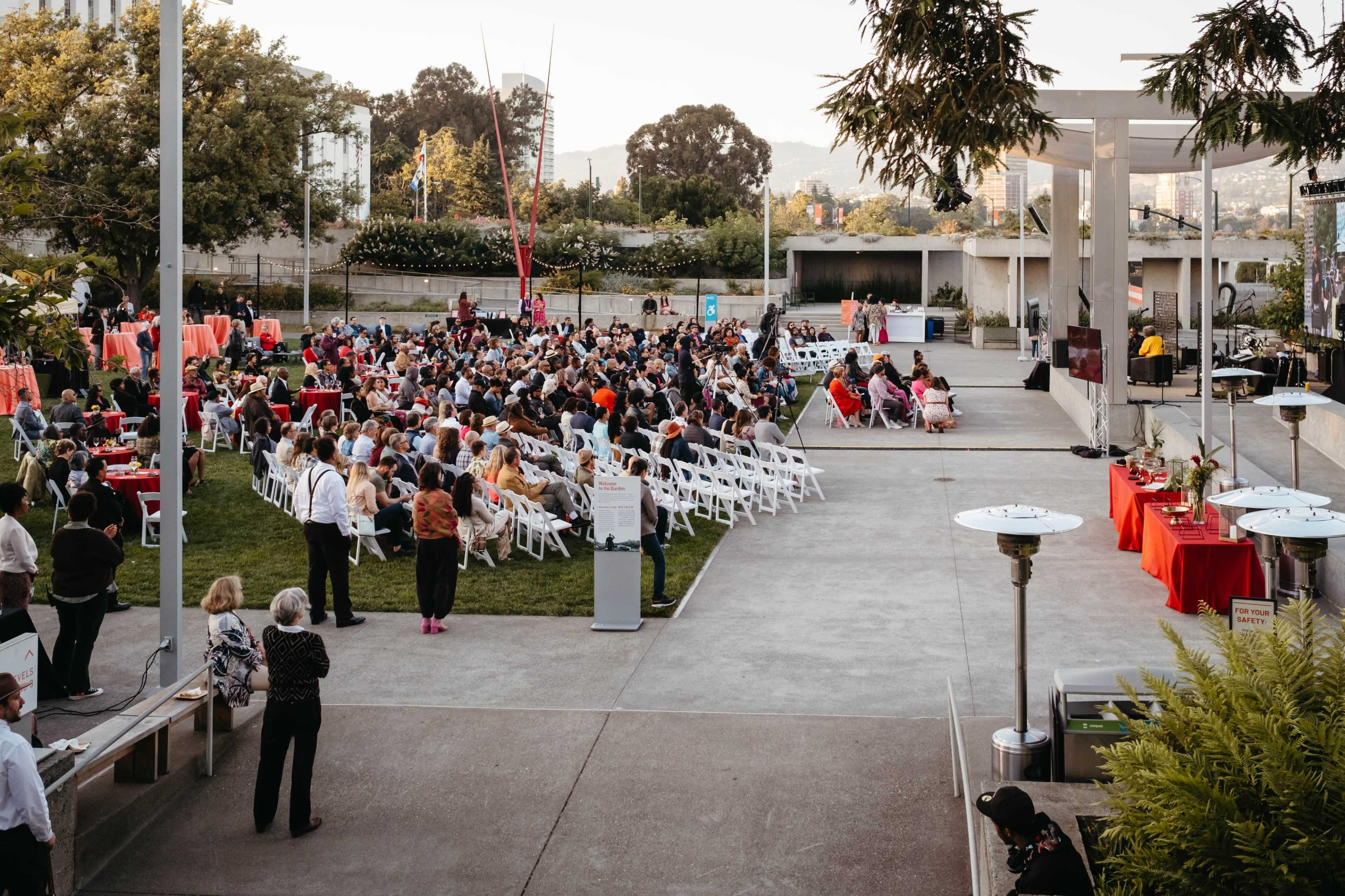 A large crowd gathers in an outdoor venue with white chairs arranged around tables, surrounded by greenery and a stage setup.