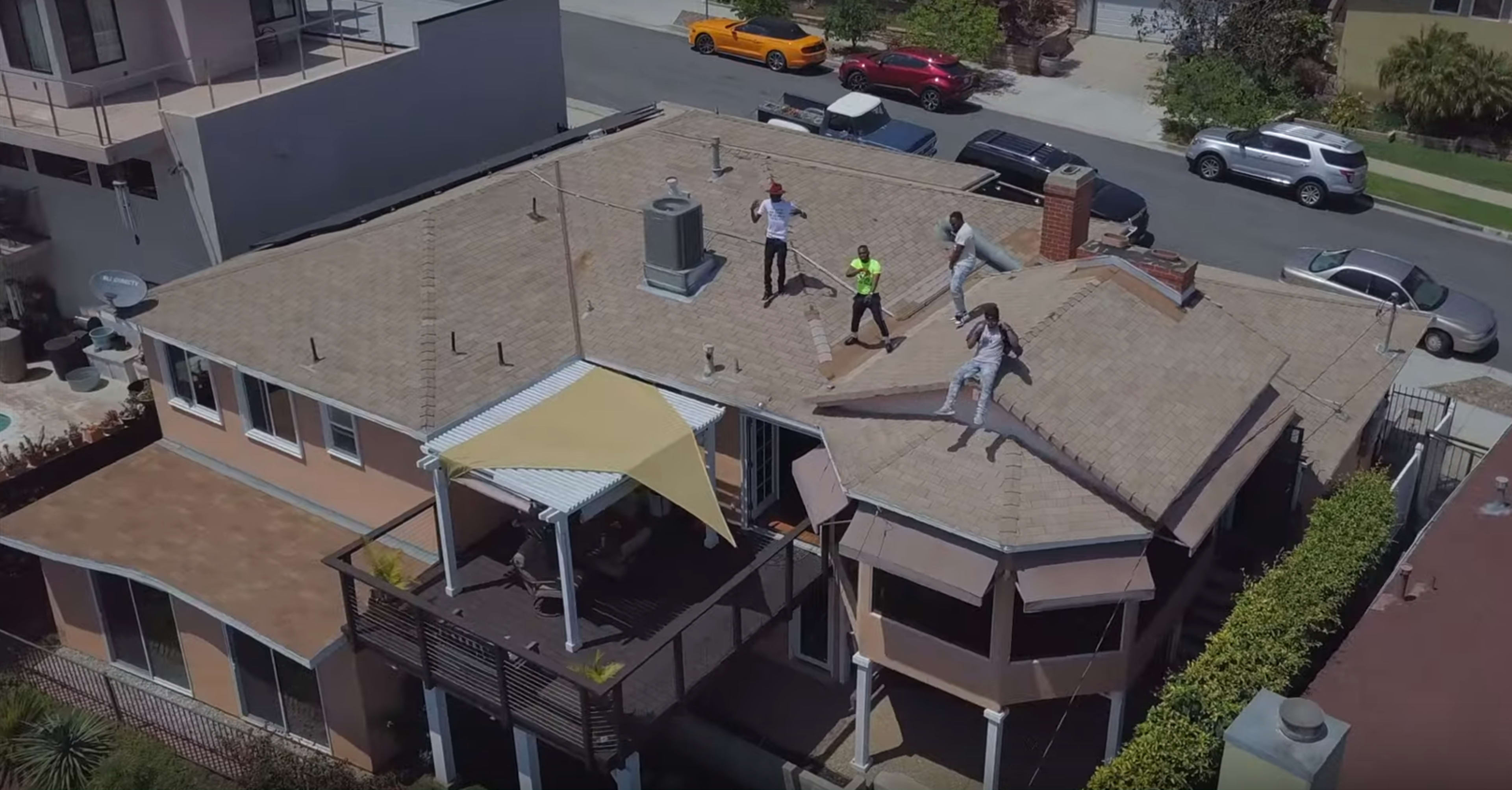 Four people are seen on the roof of a house with a deck and a large awning, while vehicles are parked on the street below.