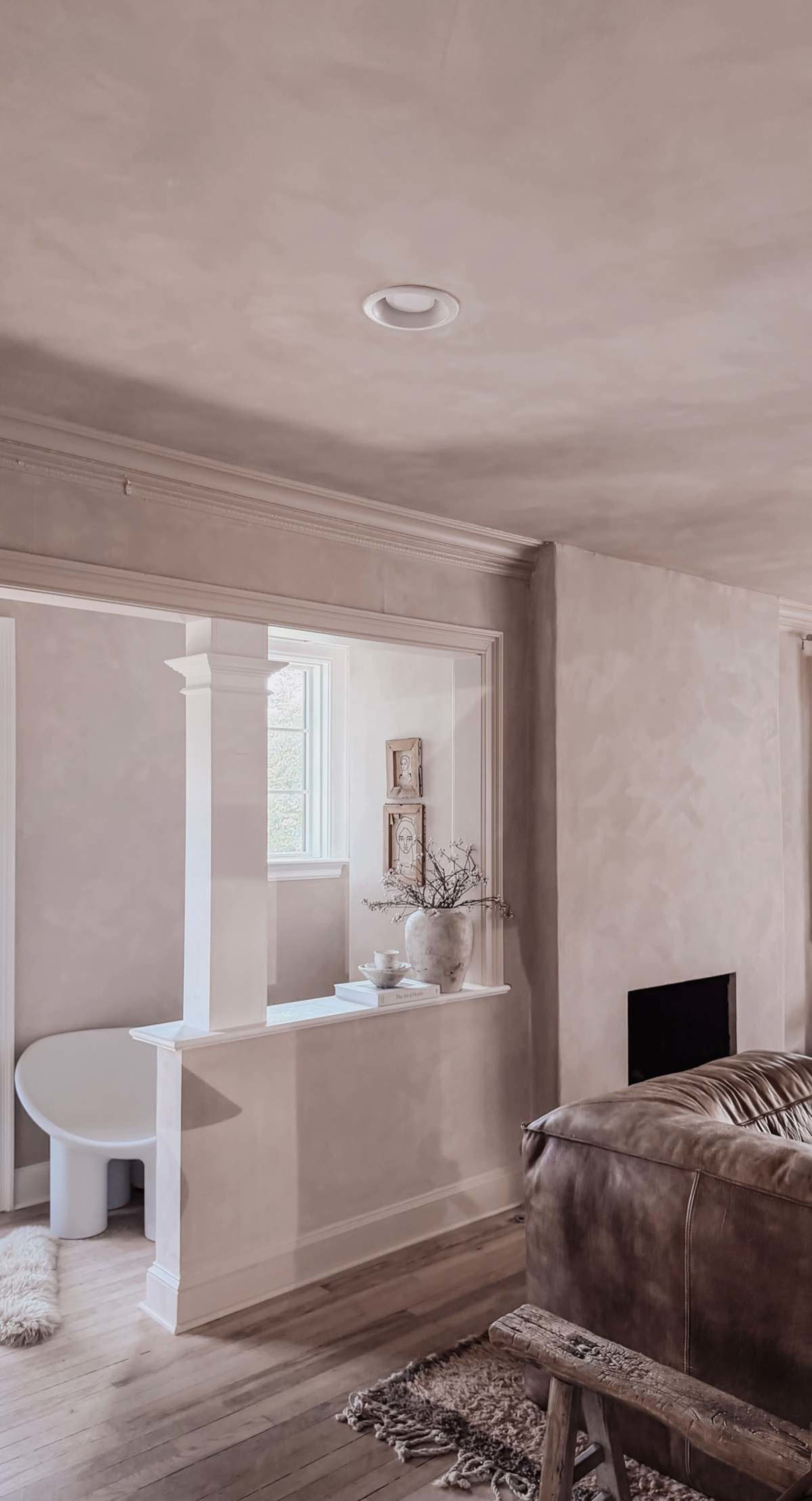The image shows a softly lit interior space featuring a minimalist design with a visible archway leading to another room, a round table, and a decorative vase on a shelf.