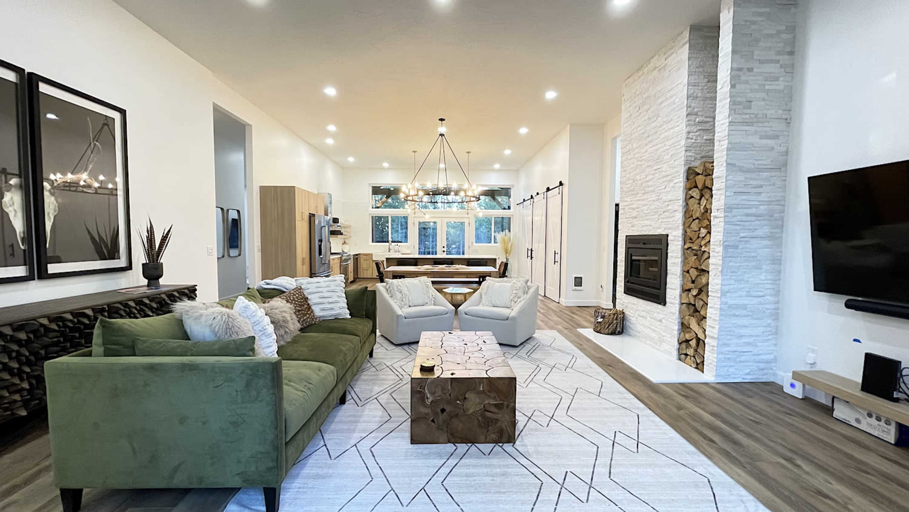 The living room features a green sofa, two armchairs, a wooden coffee table, and a fireplace surrounded by stacked wood, all arranged under a chandelier with large windows in the background.