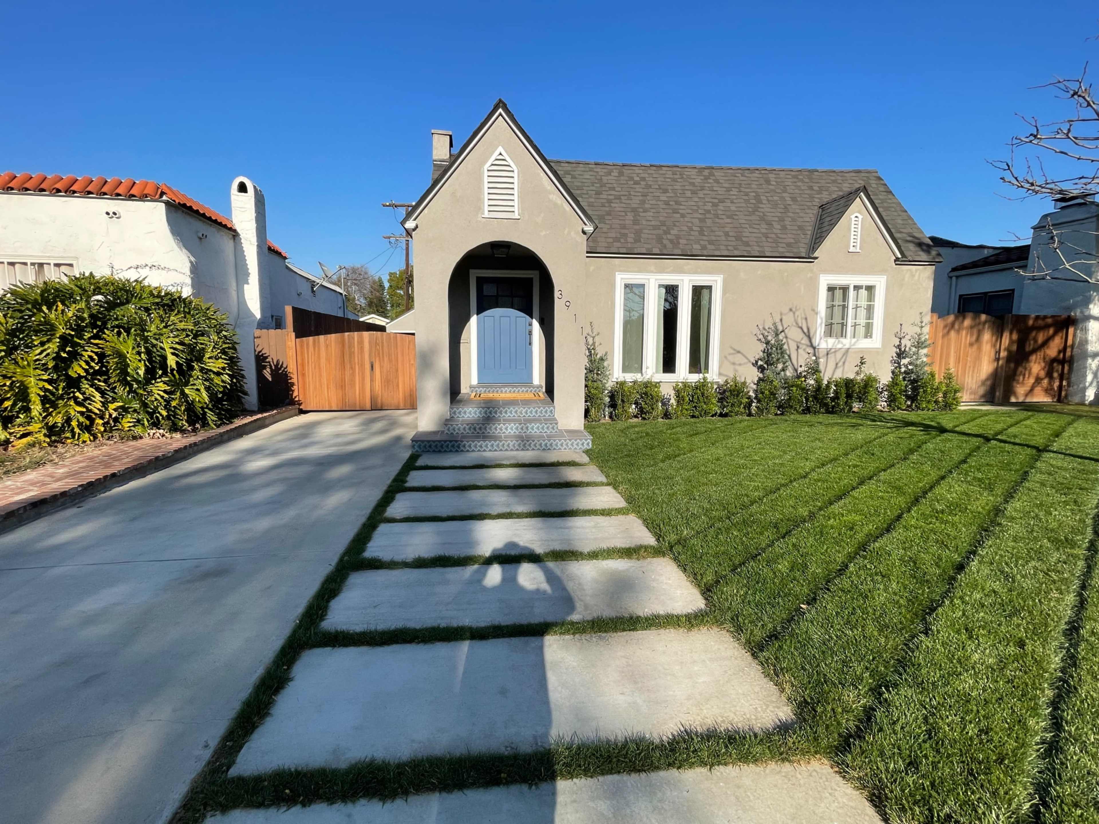 A single-story house with a gray exterior, a blue front door, and a landscaped yard featuring neatly trimmed grass and concrete pathways.