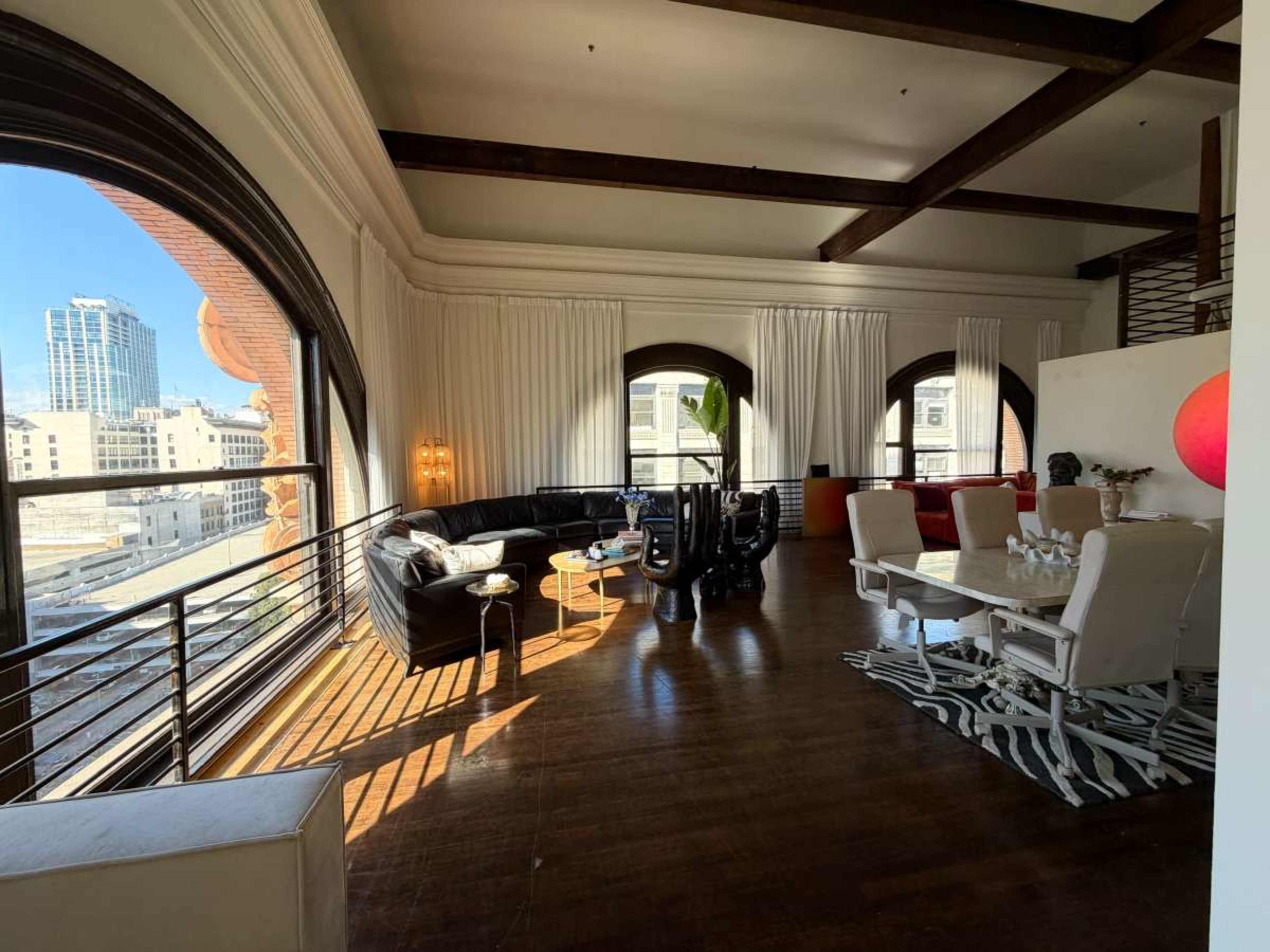 The image shows a spacious, modern living area with large windows, a black leather sofa, a white dining table, and a decorative rug on a wooden floor.