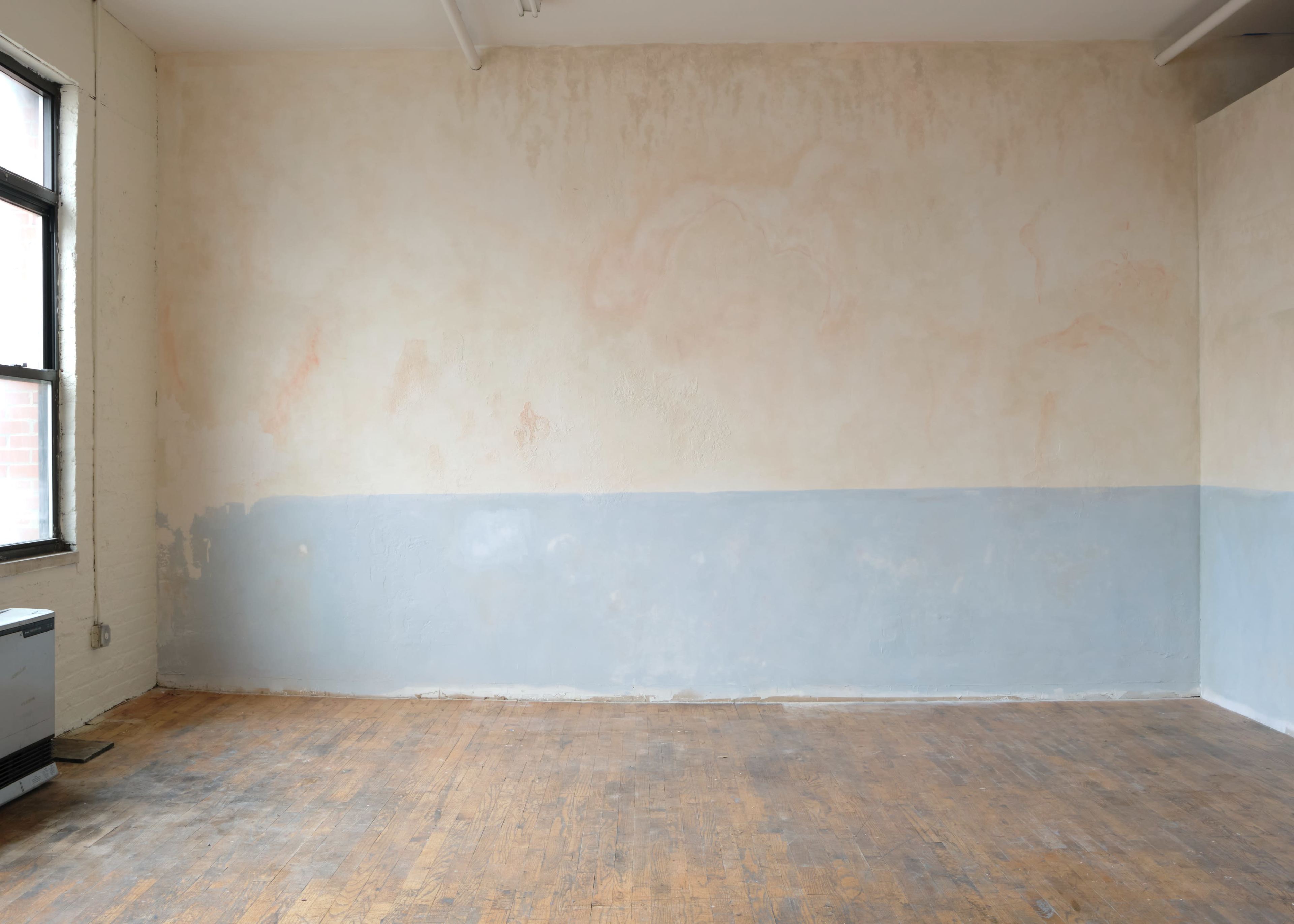 The image shows a minimalist room with a partially painted wall featuring a soft color gradient and a wooden floor.