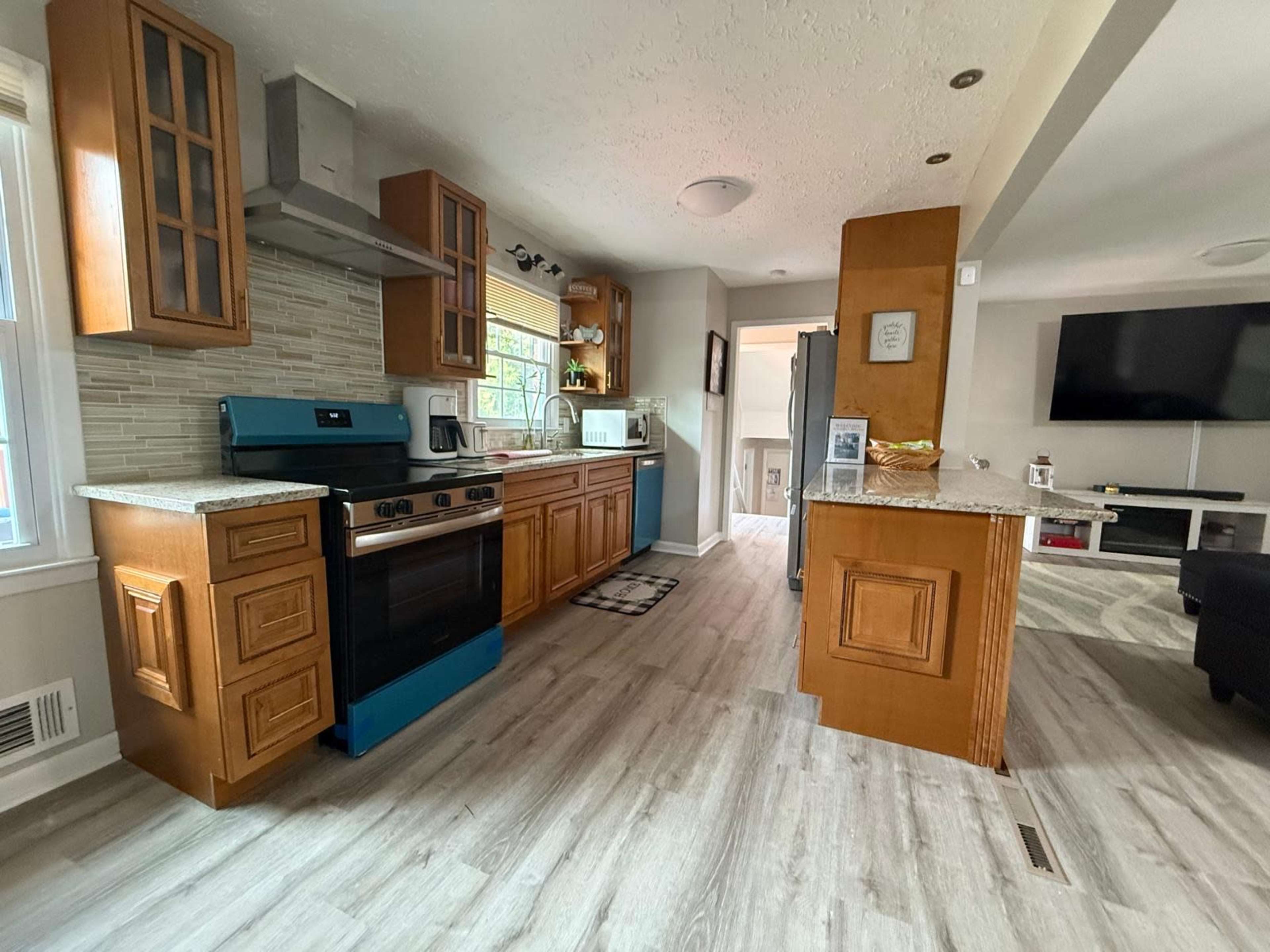 The image shows a modern kitchen featuring wooden cabinetry, a blue stovetop, granite countertops, and an open layout connected to a living area.