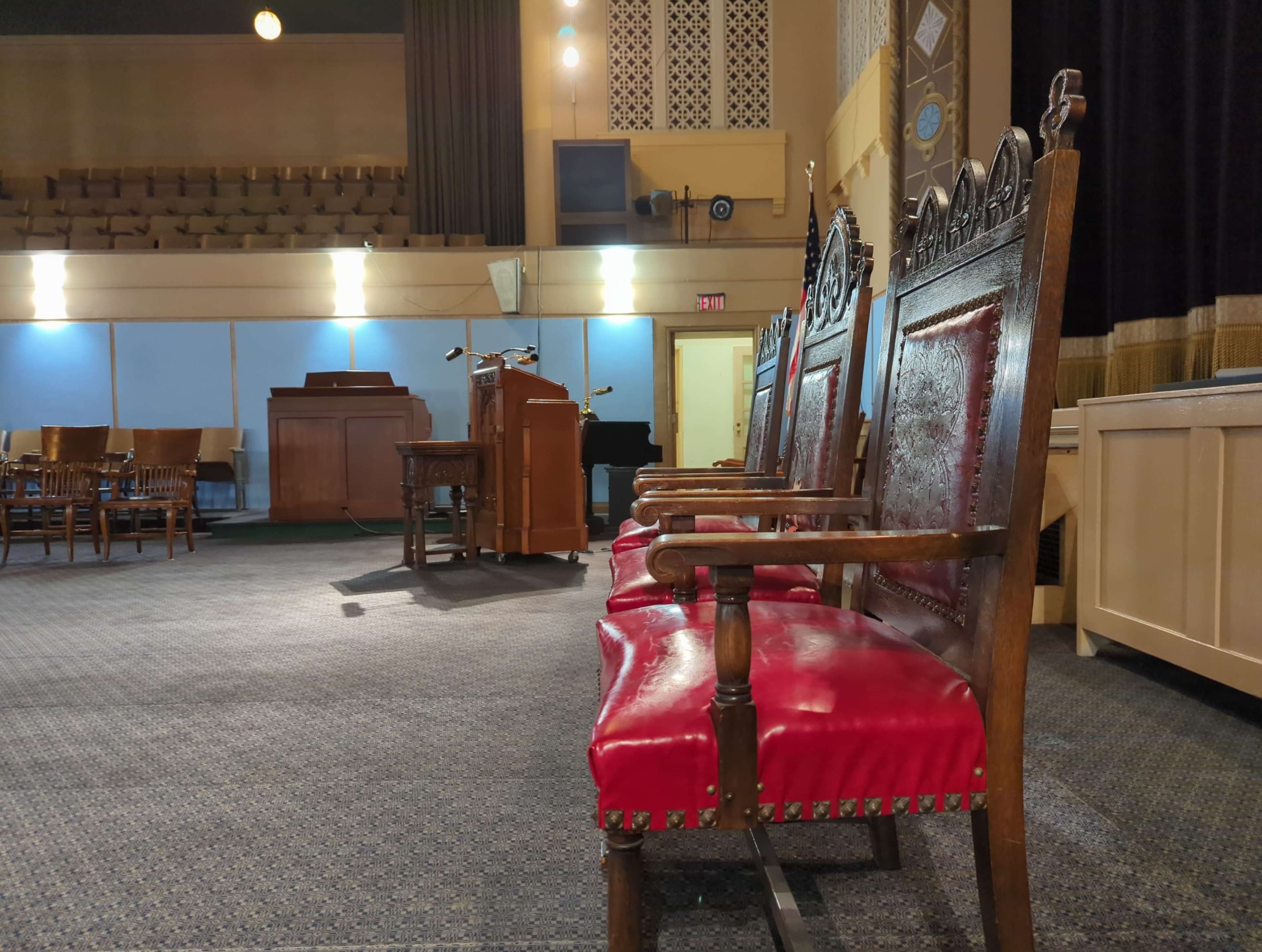 The image shows a row of ornate wooden chairs with red upholstery positioned in a theater or auditorium setting, facing a wooden lectern and piano in the background.