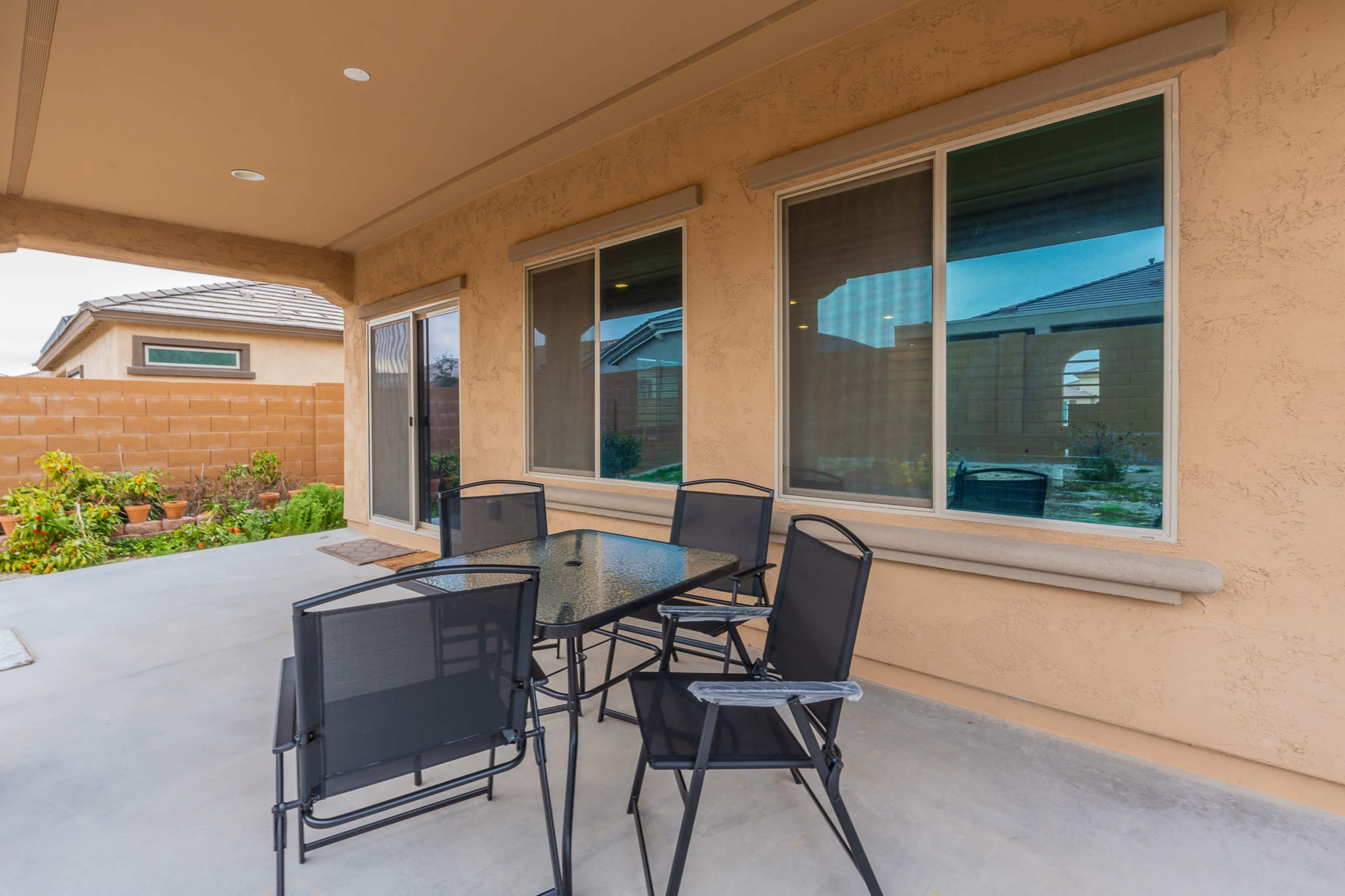 The image shows a patio area with a glass-top table and several black chairs, set against a backdrop of a beige stucco wall and greenery.