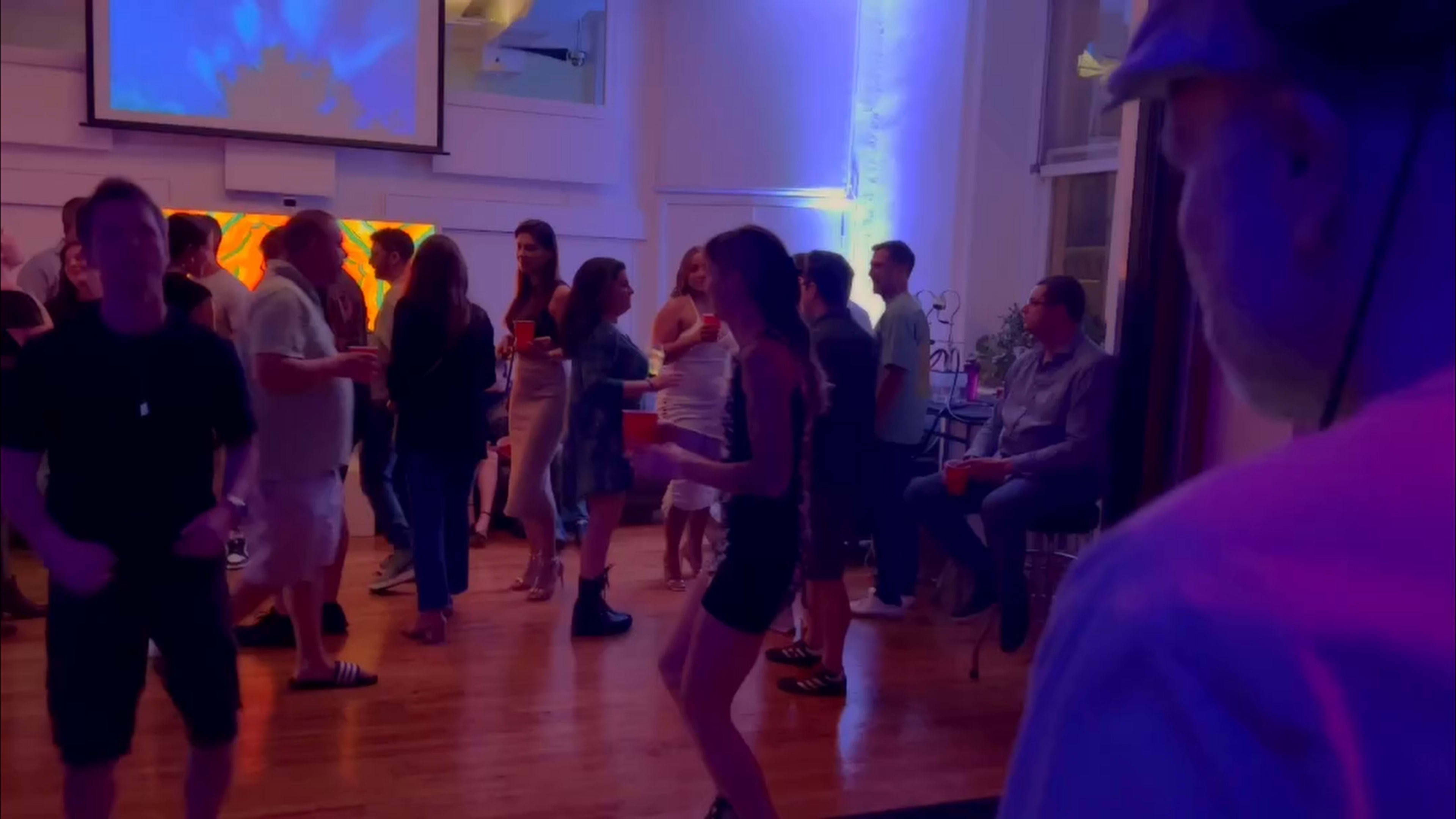 A group of people is dancing and socializing in a well-lit indoor space with colorful lighting.