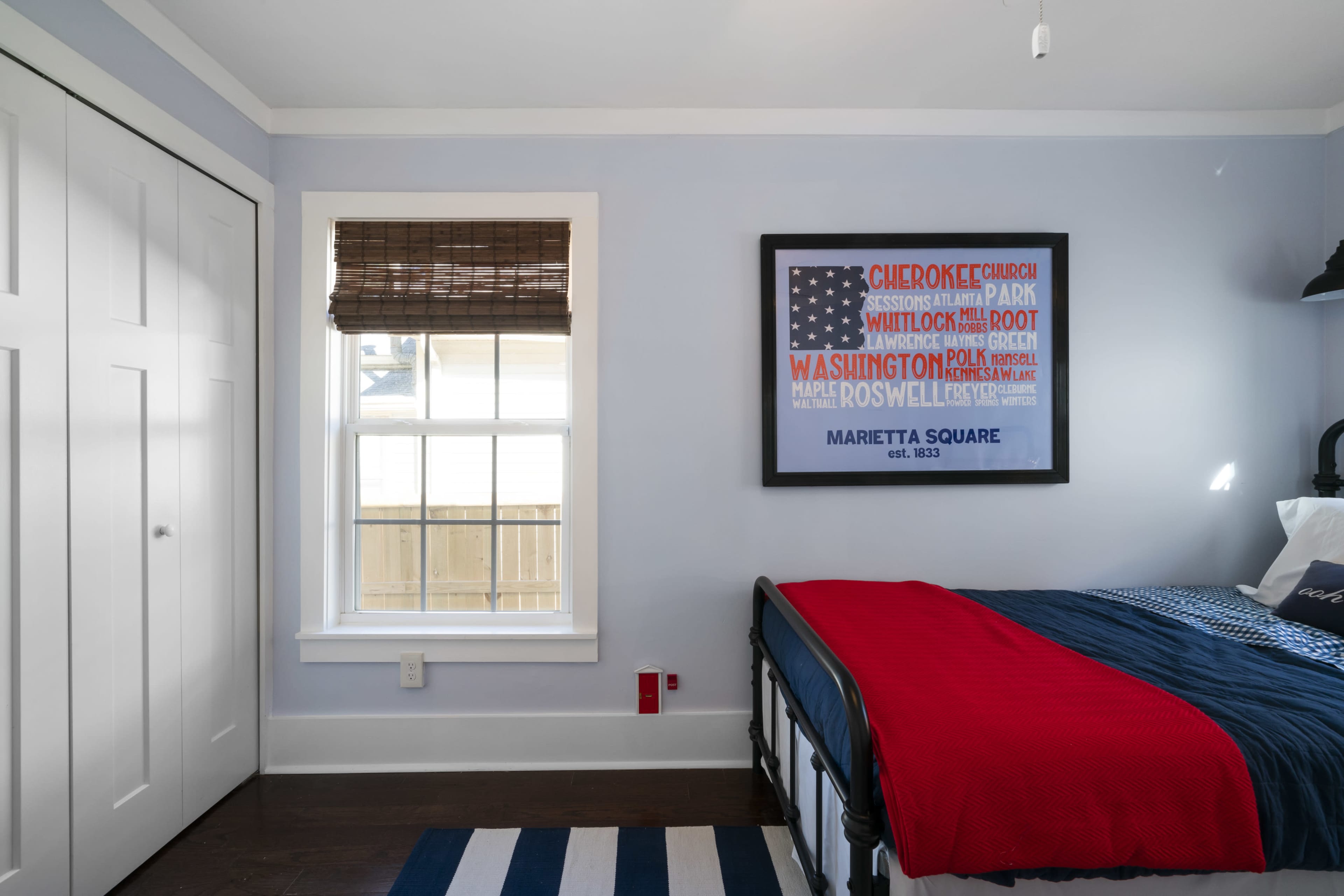A neatly organized bedroom features a bed with a blue and red cover, a window with a wooden blind, and a framed artwork on the wall.