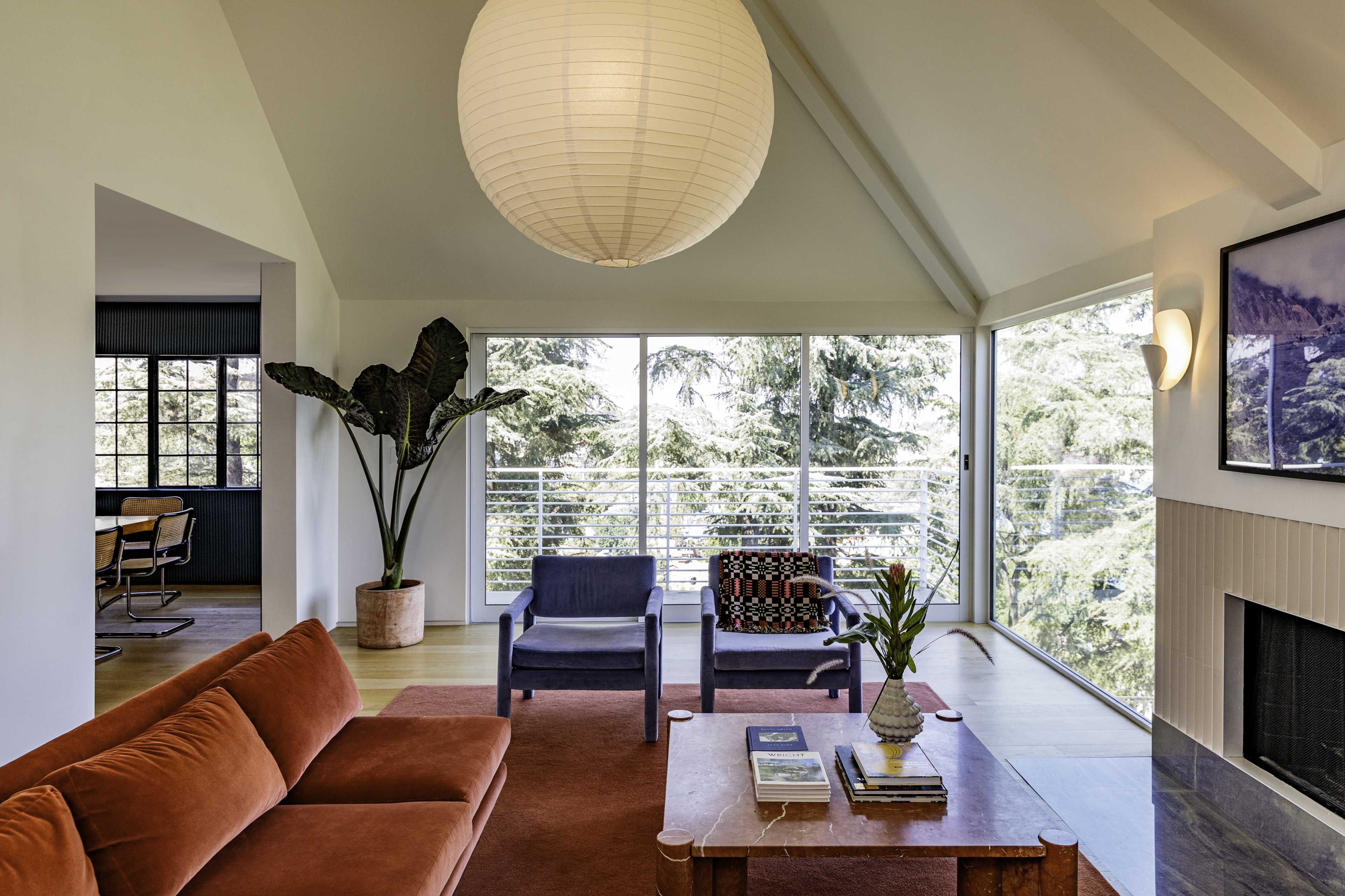 A modern living room with a high ceiling, large windows, and a mid-century sofa, featuring a coffee table and potted plants.