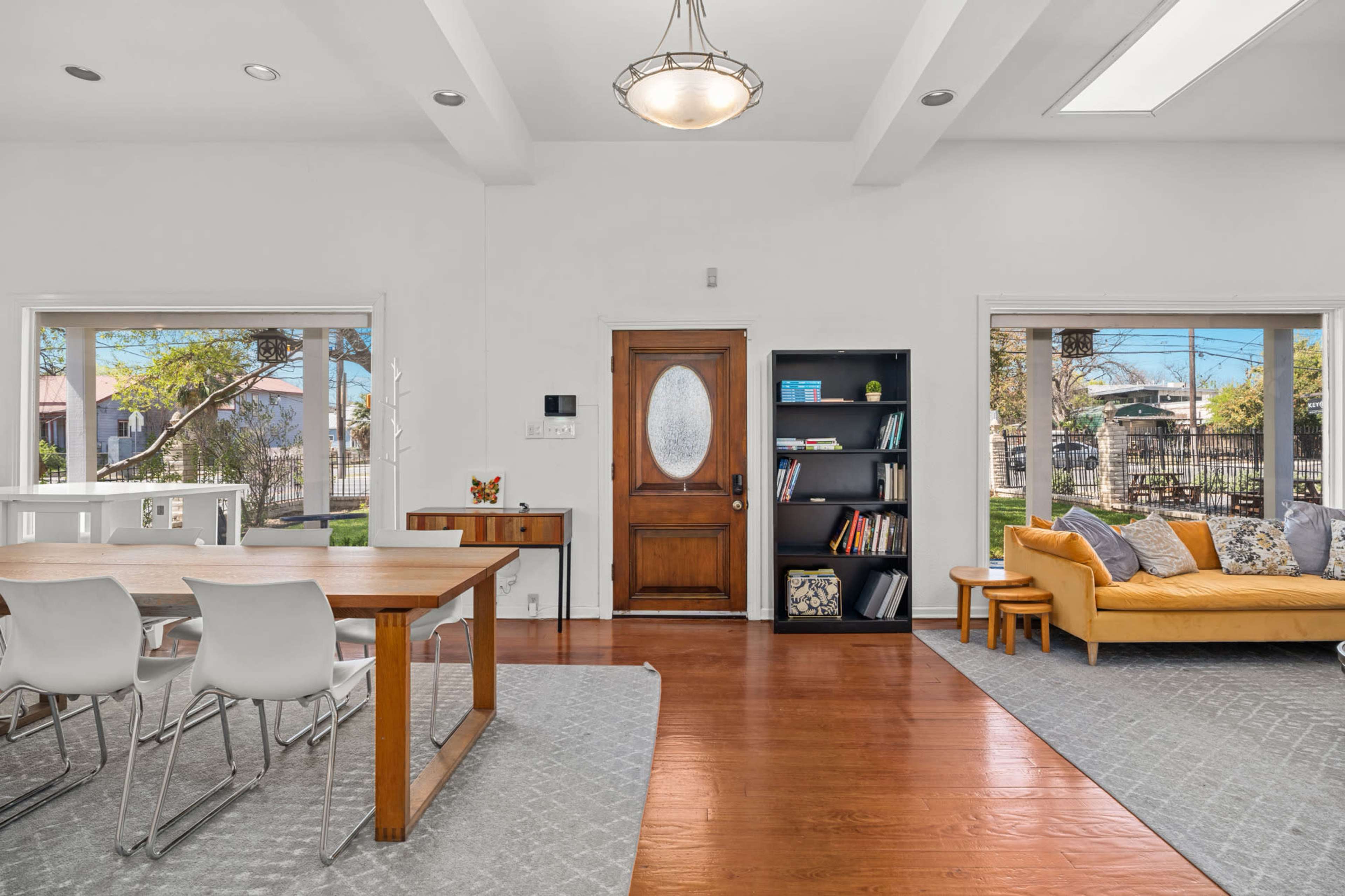 The image shows a bright and open living space featuring a dining area with a wooden table and chairs, a cozy seating area with a yellow couch, and large windows allowing natural light.