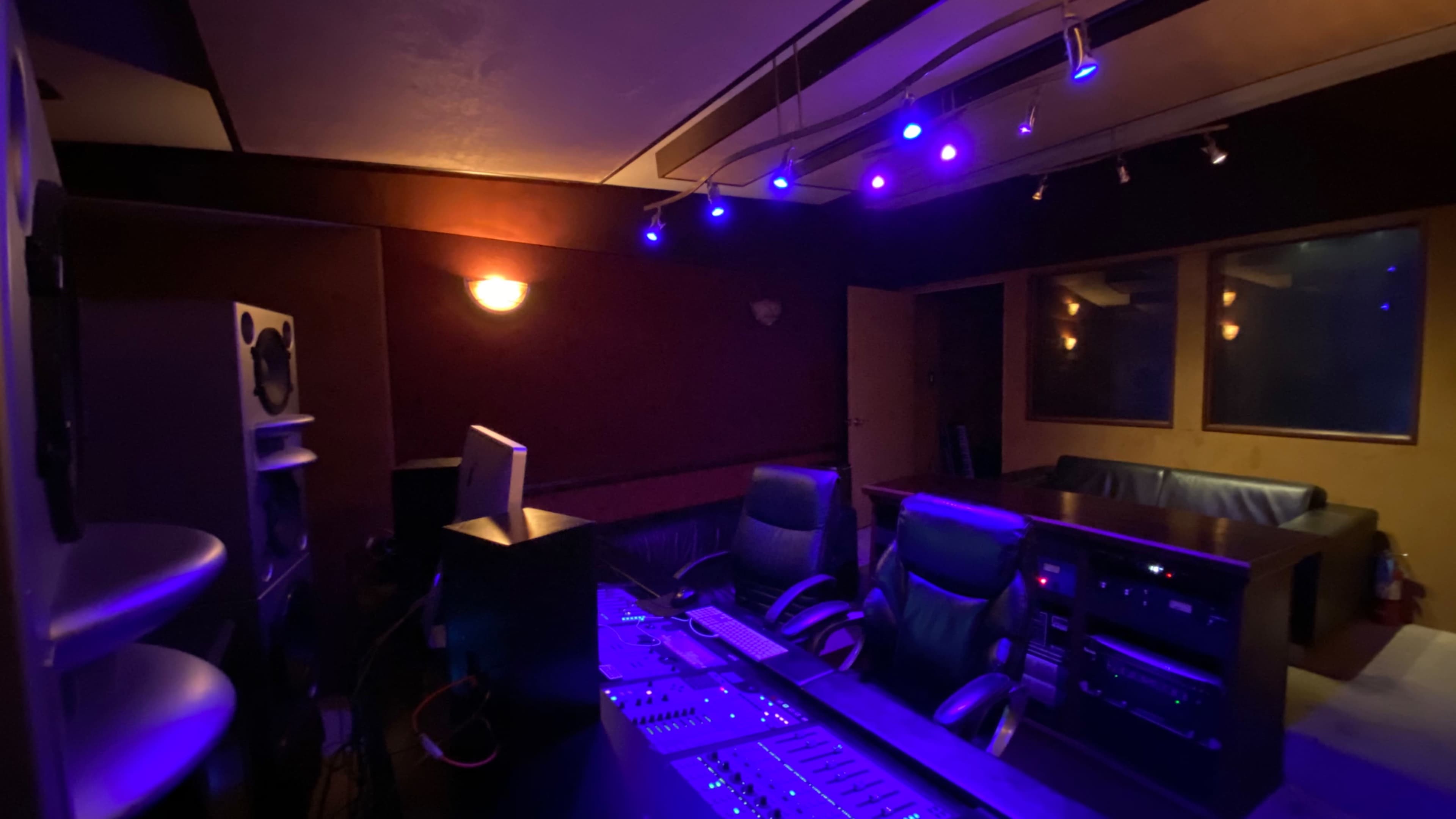 The image shows a dimly lit recording studio featuring a sound mixing desk, several audio monitors, and comfortable seating areas.