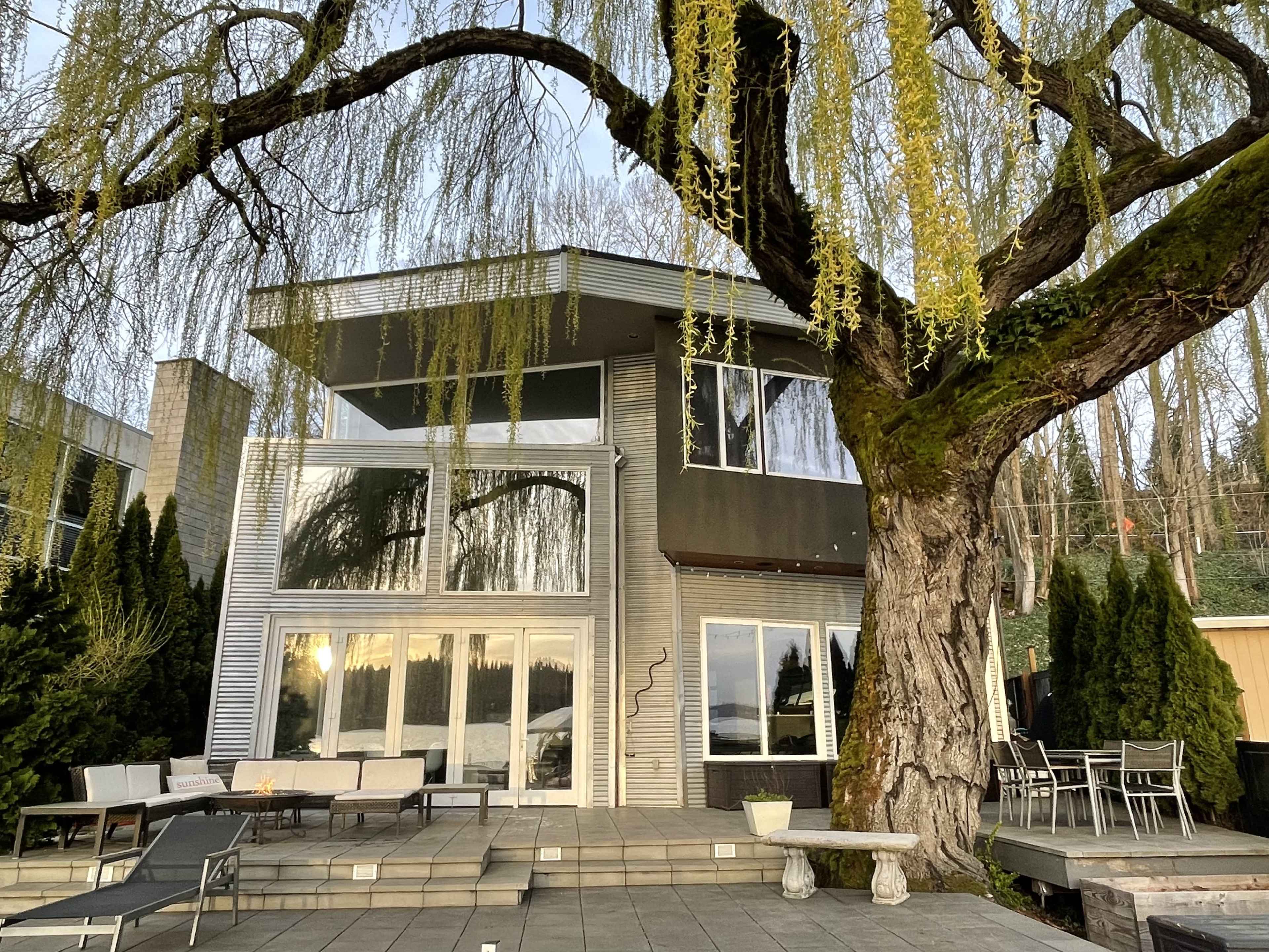 A modern two-story house with large windows is situated next to a large willow tree, surrounded by a patio and various outdoor seating areas.