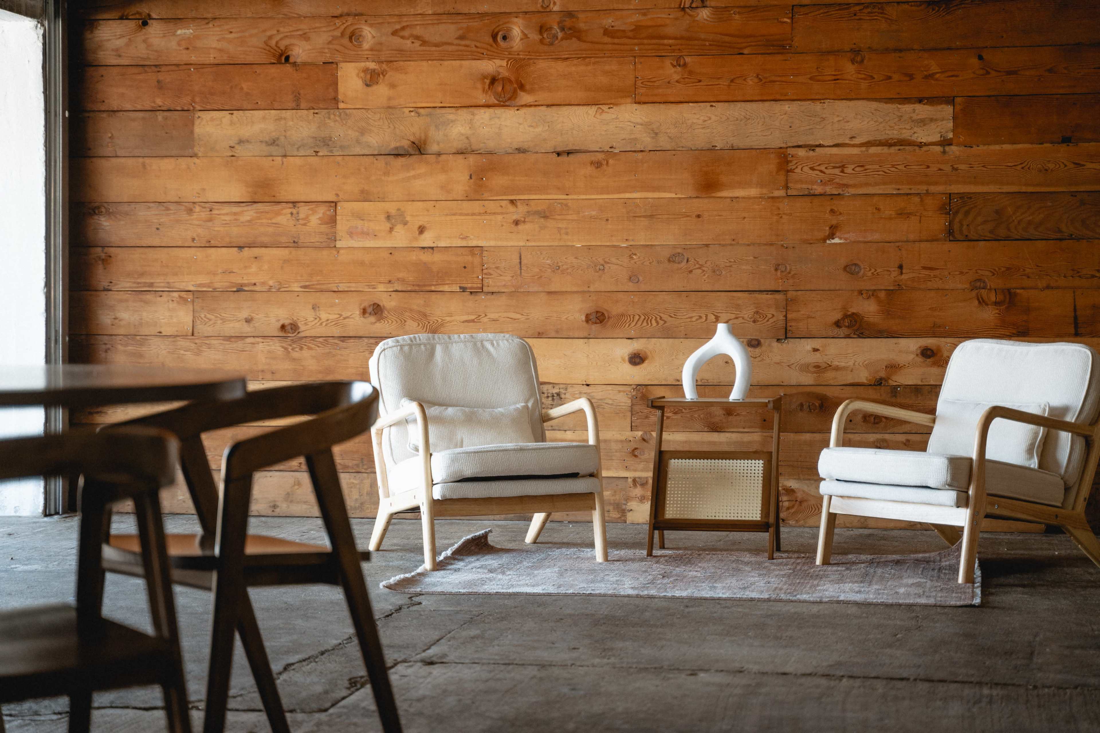 The image depicts a cozy seating area with two upholstered chairs, a small wooden table, and a vase on top, set against a rustic wooden wall.