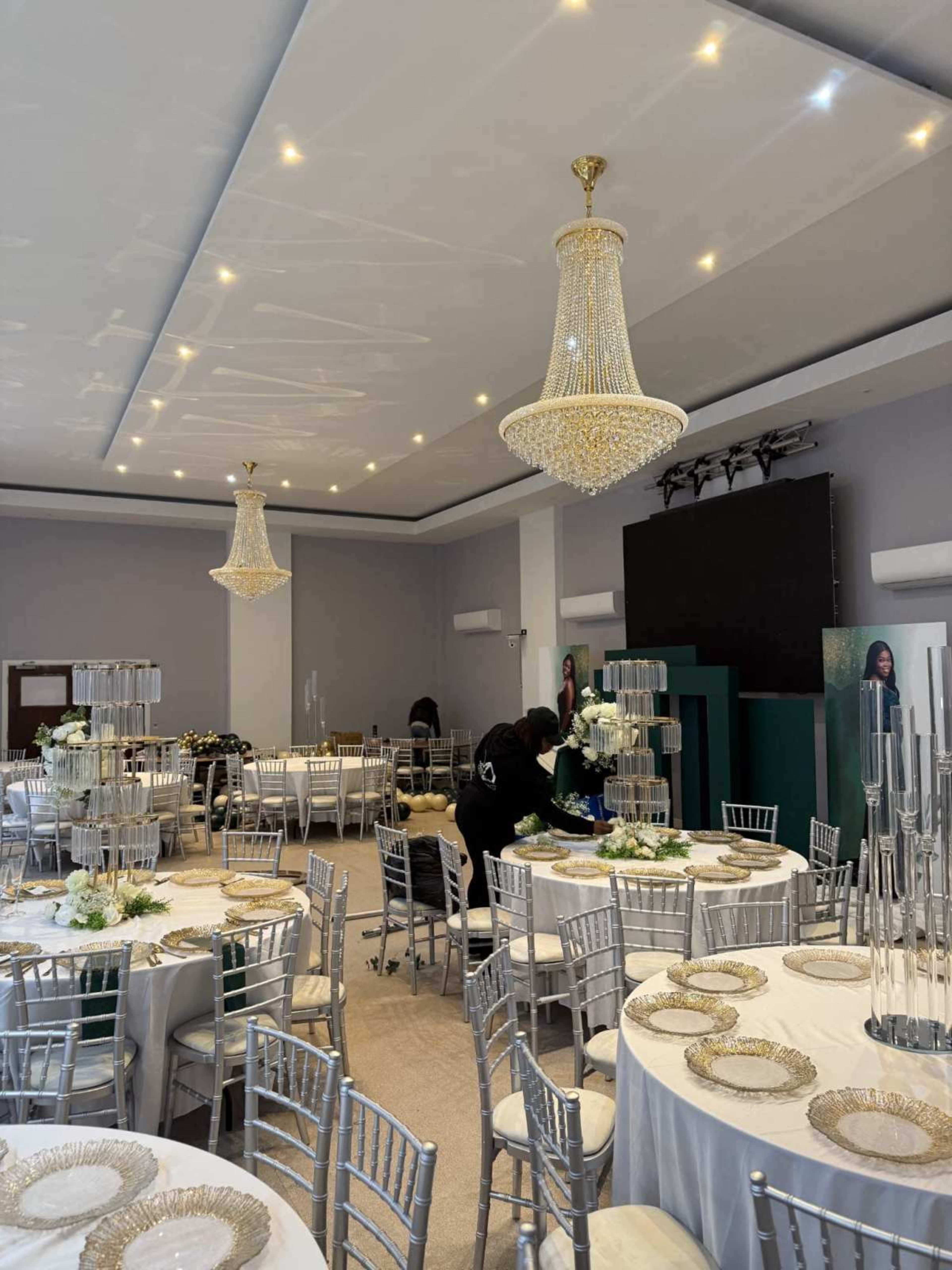 The image shows a decorated event space with round tables set for a formal gathering, featuring elegant chandeliers and a staff member preparing the area.