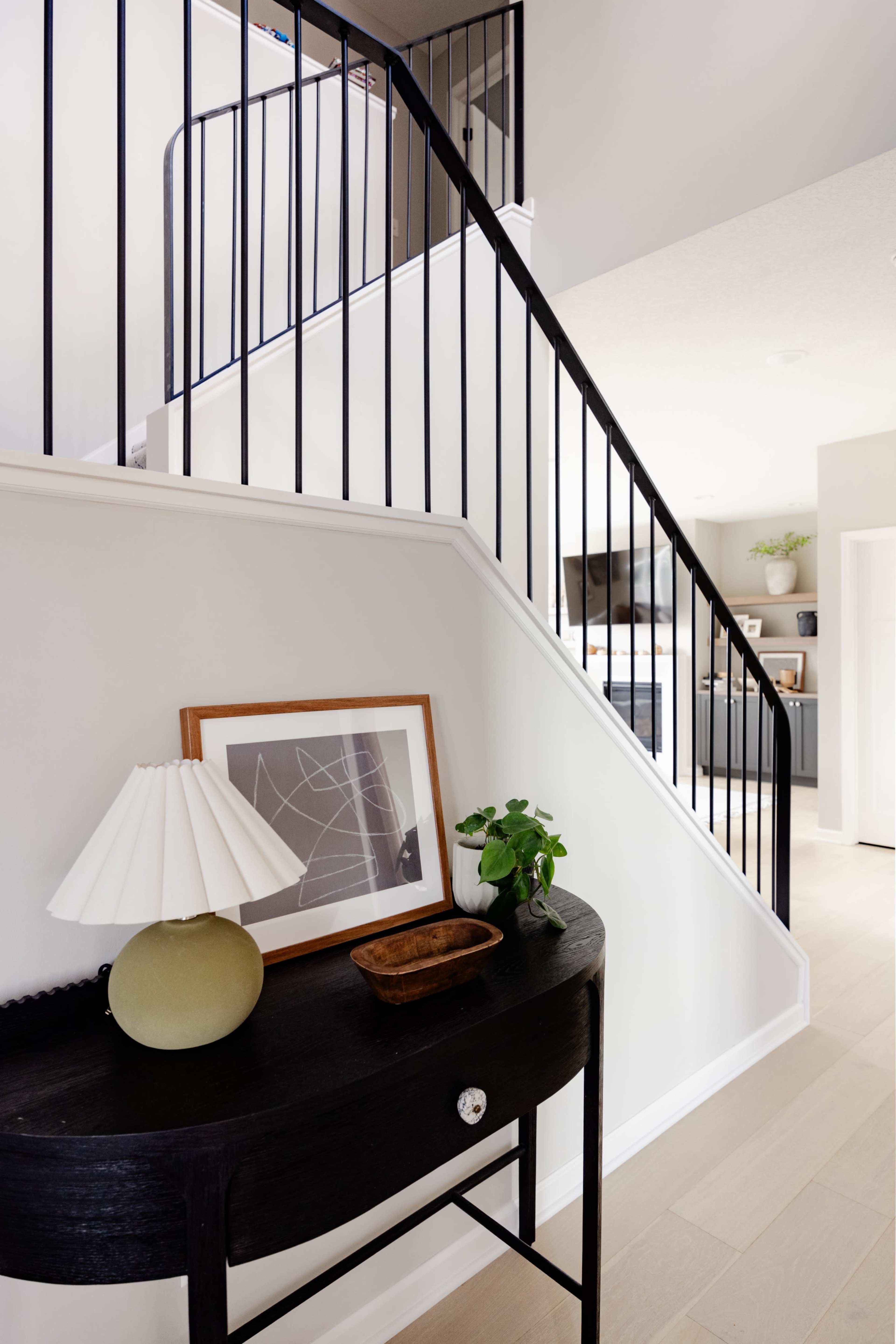 The image shows a well-lit entryway featuring a black console table with a lamp, a framed artwork, and a small plant, alongside a staircase with black railing.