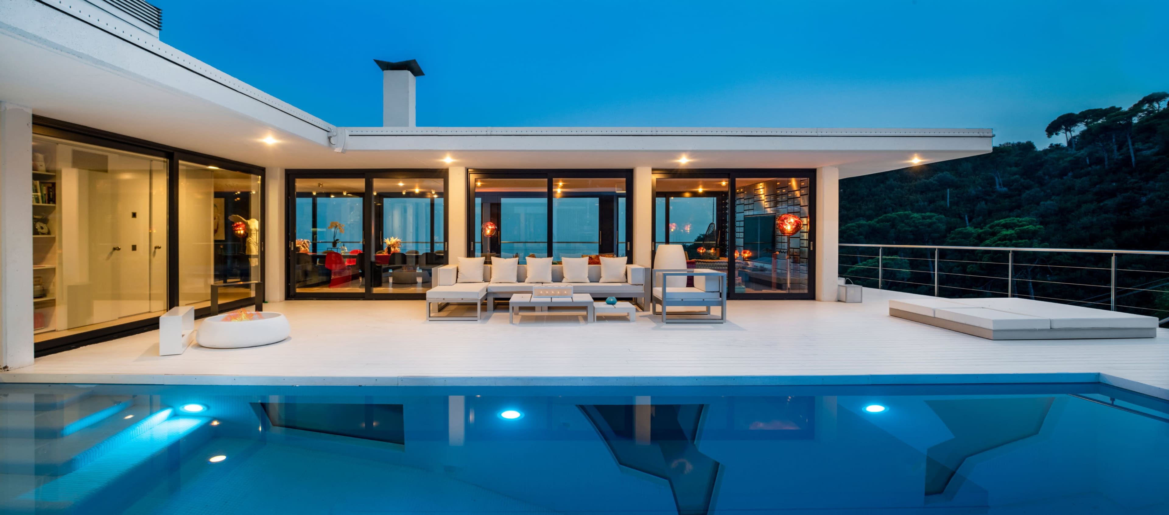 A modern villa features a spacious deck with seating and a swimming pool, overlooking a wooded area at dusk.