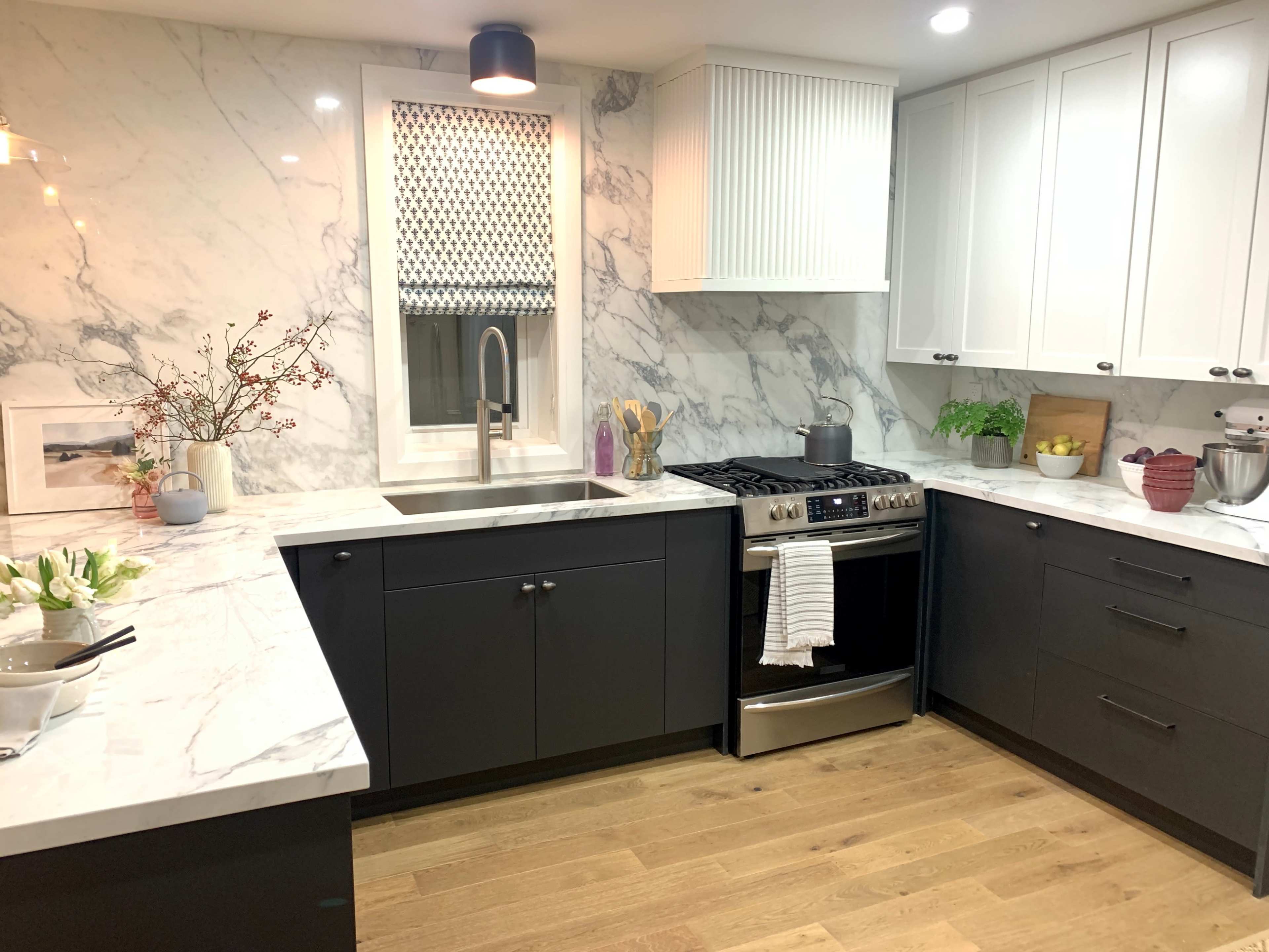 A modern kitchen features white and gray marble countertops, dark cabinetry, and stainless steel appliances.