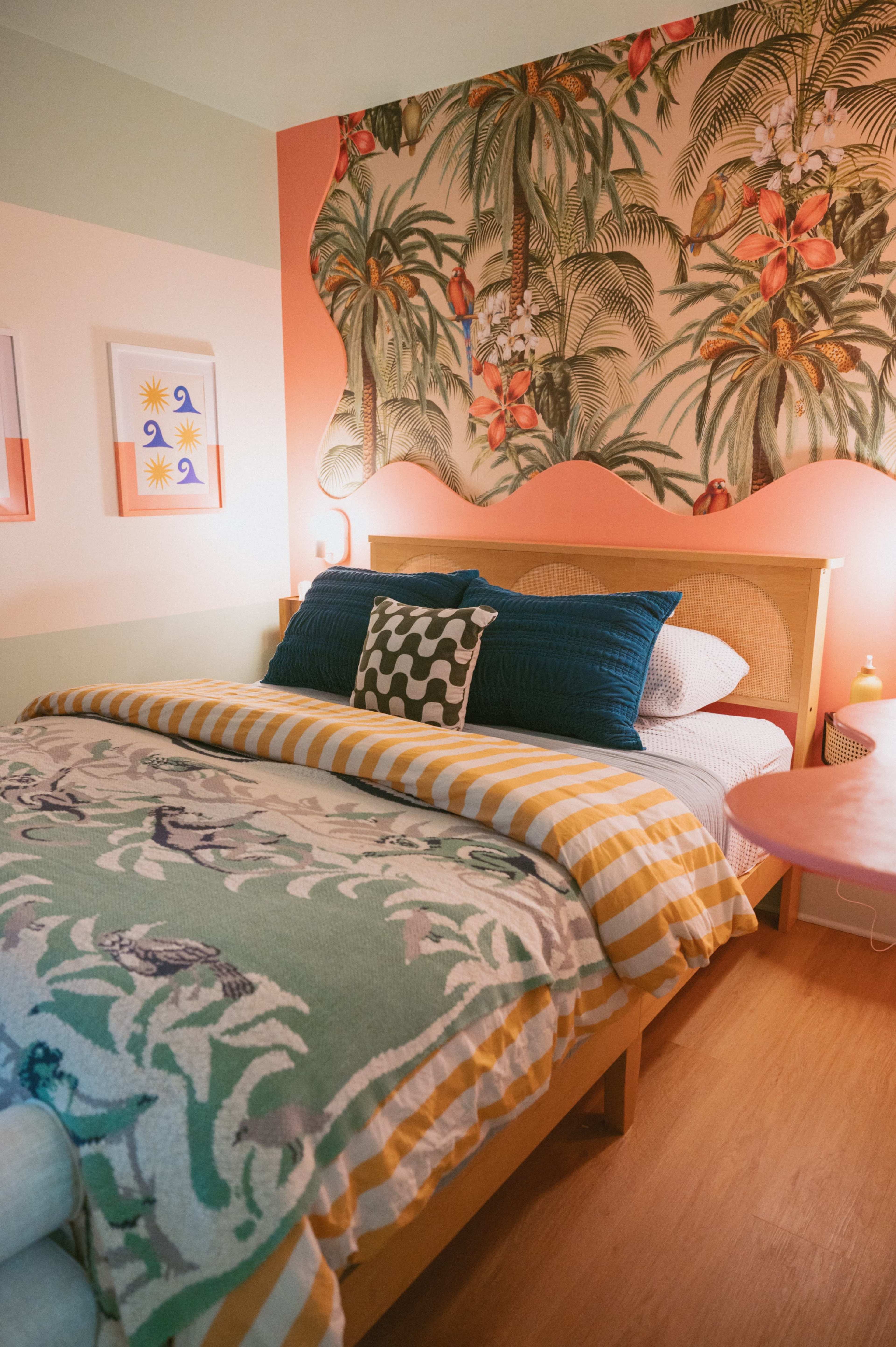 A bedroom features a bed with patterned bedding, a tropical wallpaper backdrop, and colorful decor elements.