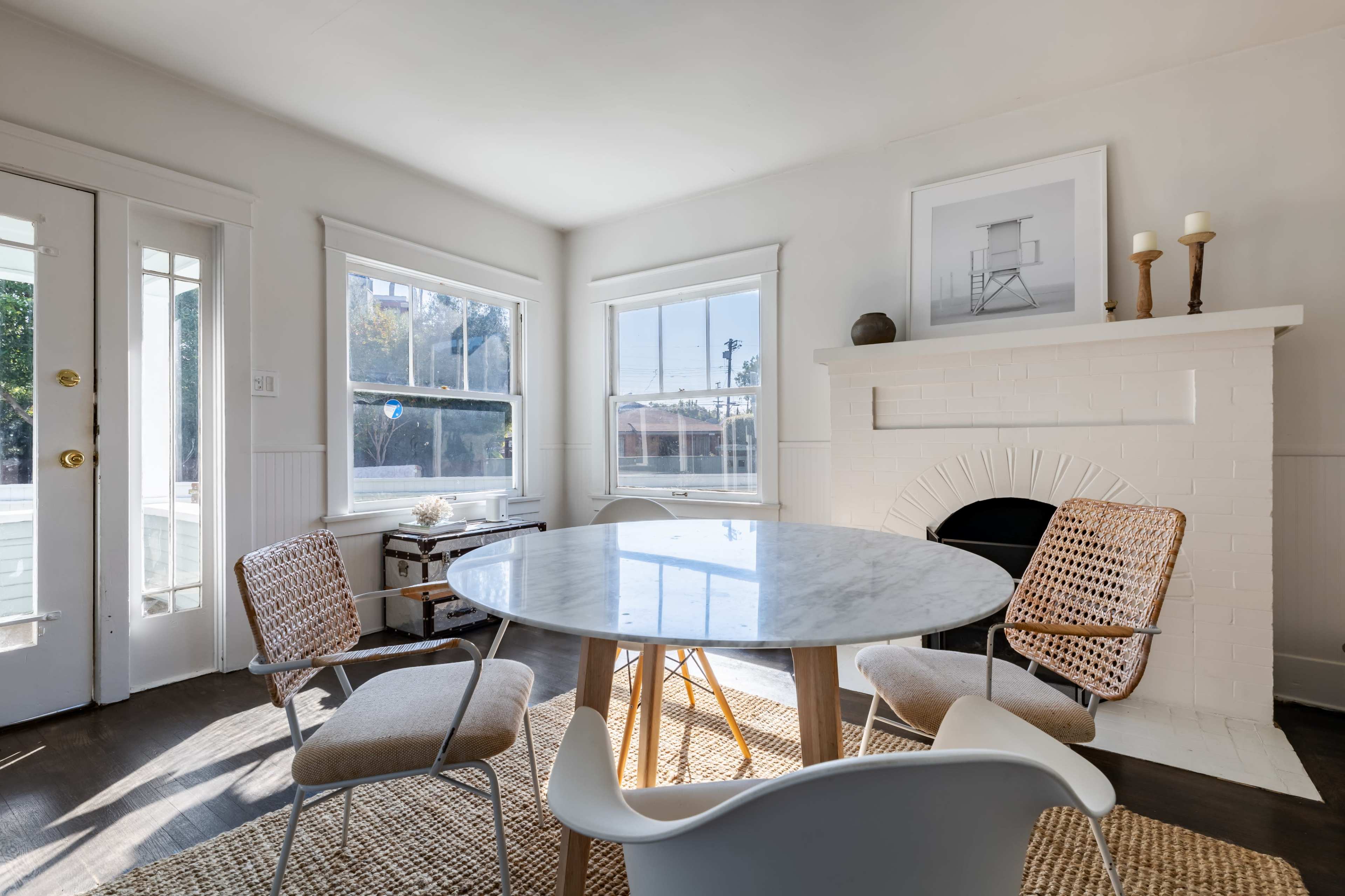A bright dining area features a round marble table surrounded by woven chairs and a white brick fireplace with candles.