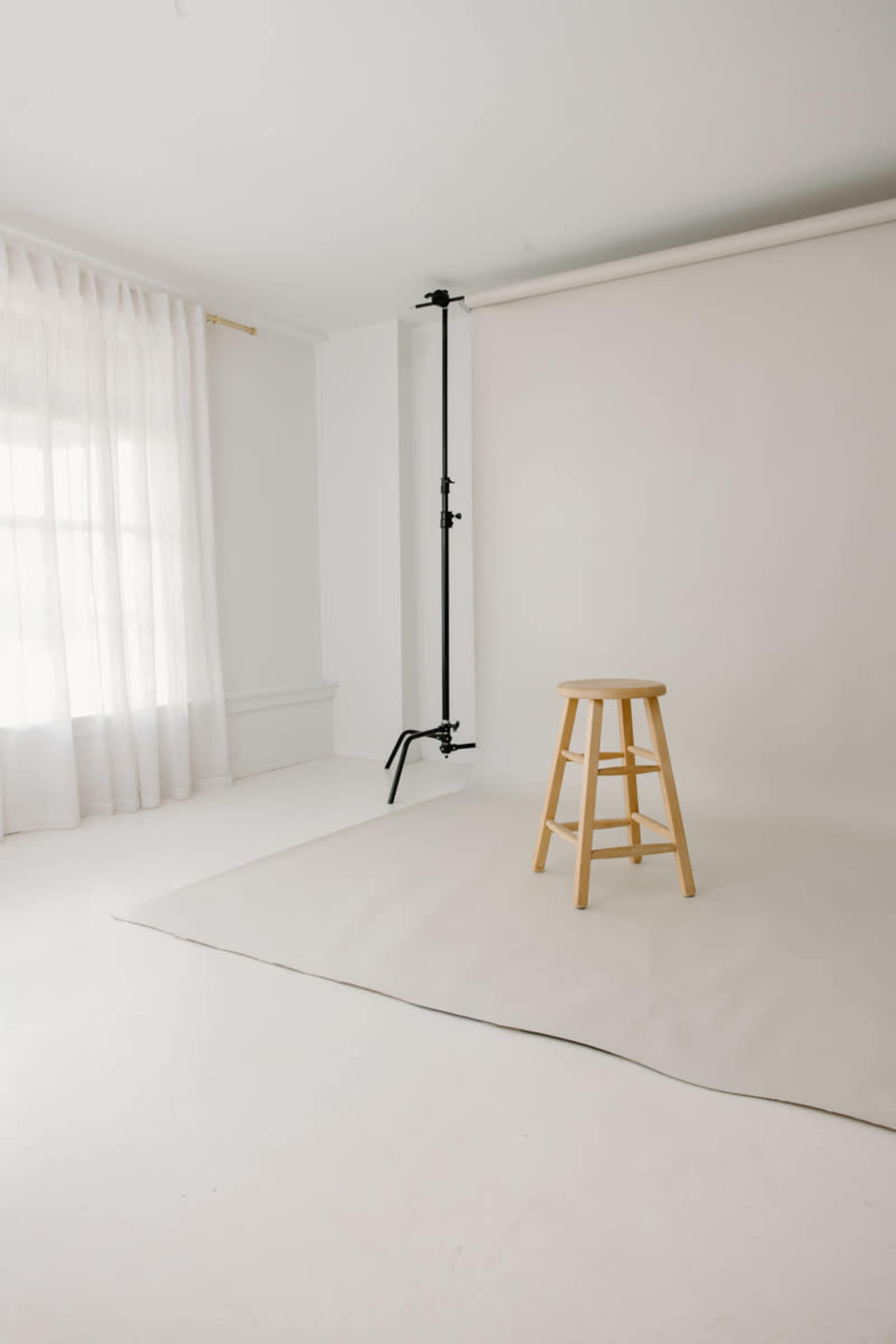 A wooden stool stands alone on a light-colored floor in a bright, minimalistic studio with sheer curtains and a plain backdrop.