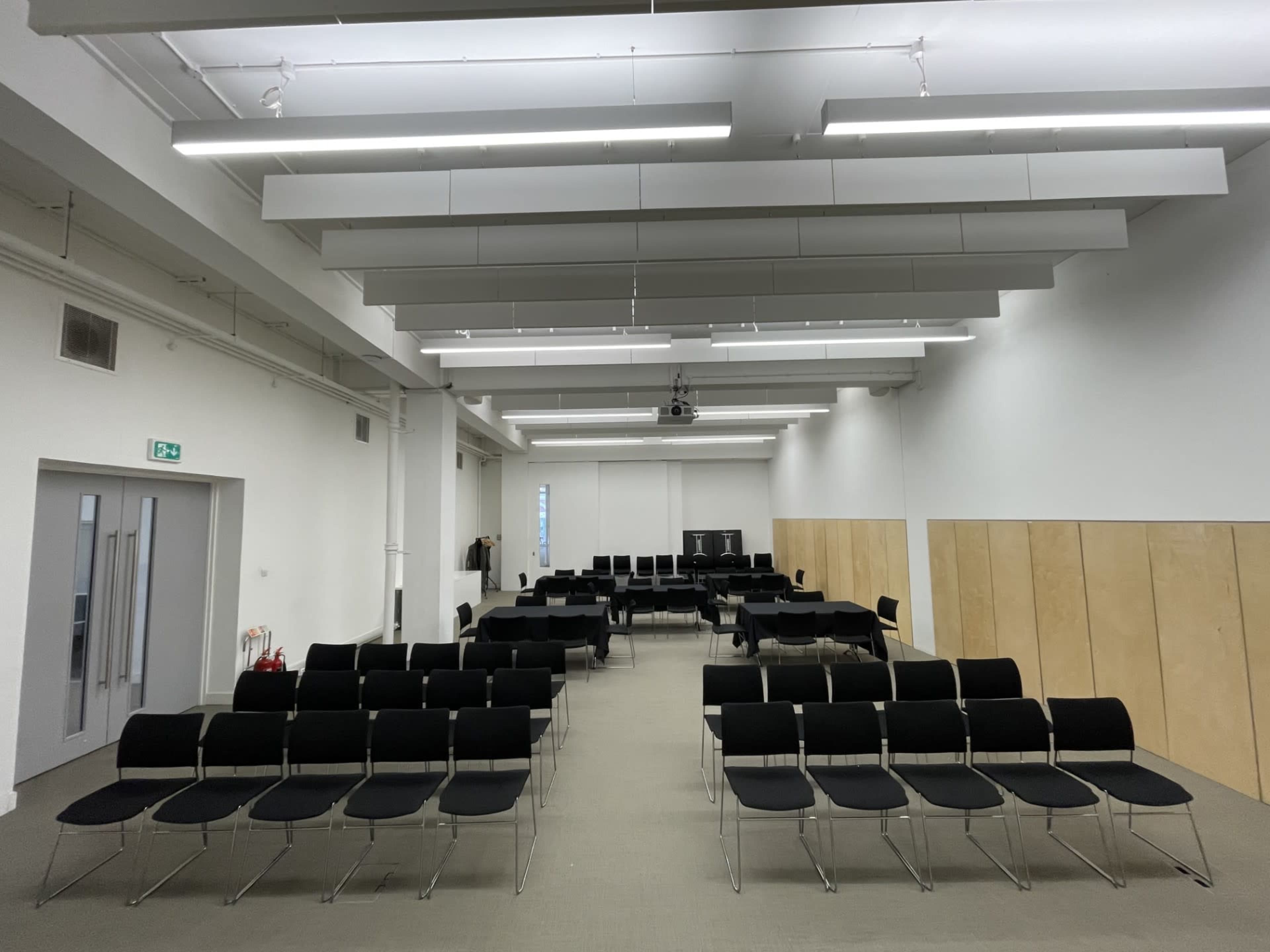 The image shows a spacious room set up with rows of black chairs facing a projection area, featuring white walls and a gray floor.
