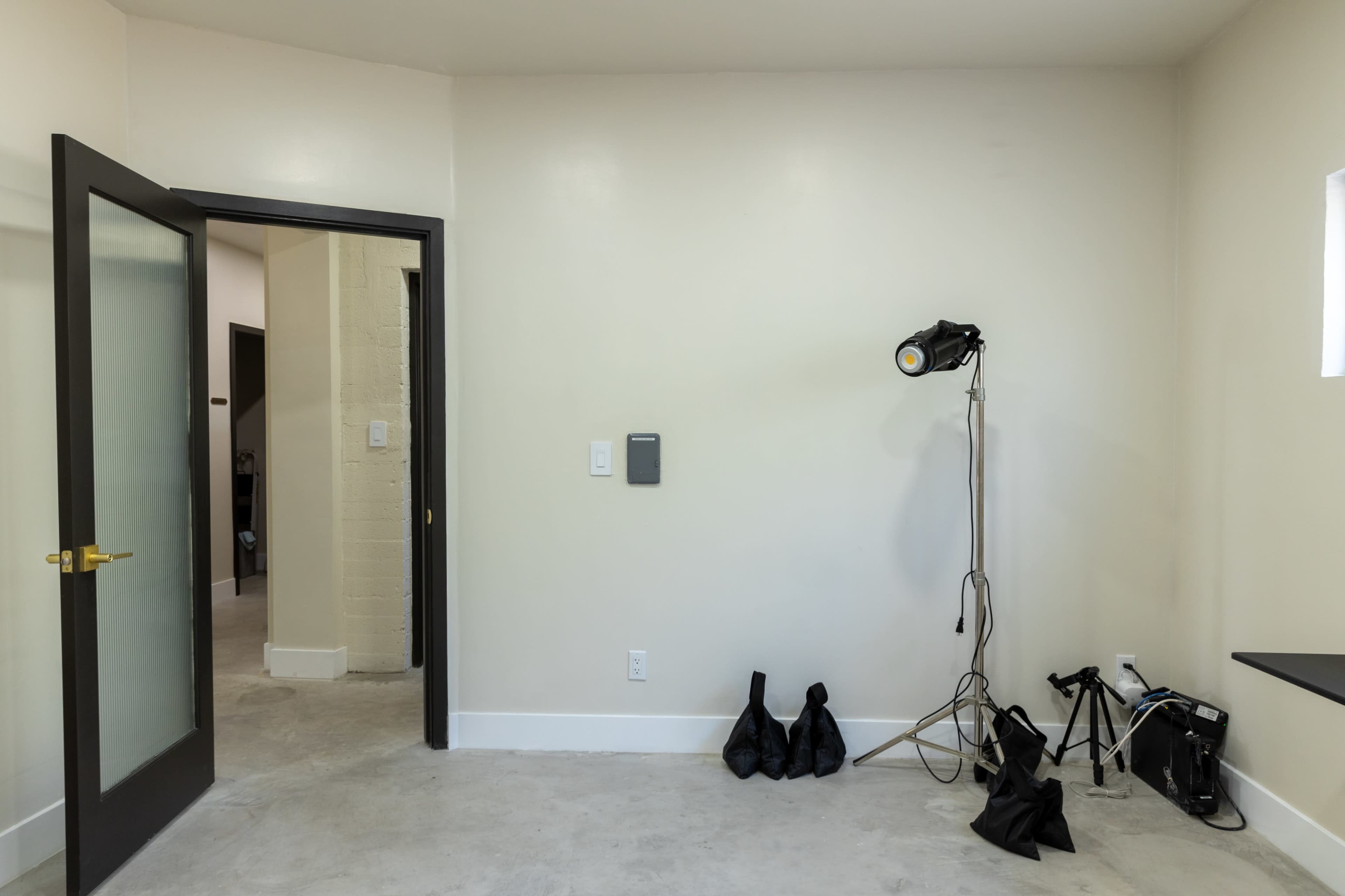 A minimalistic room with a gray floor, a door leading to another space, and a light stand next to two black bags and camera equipment.