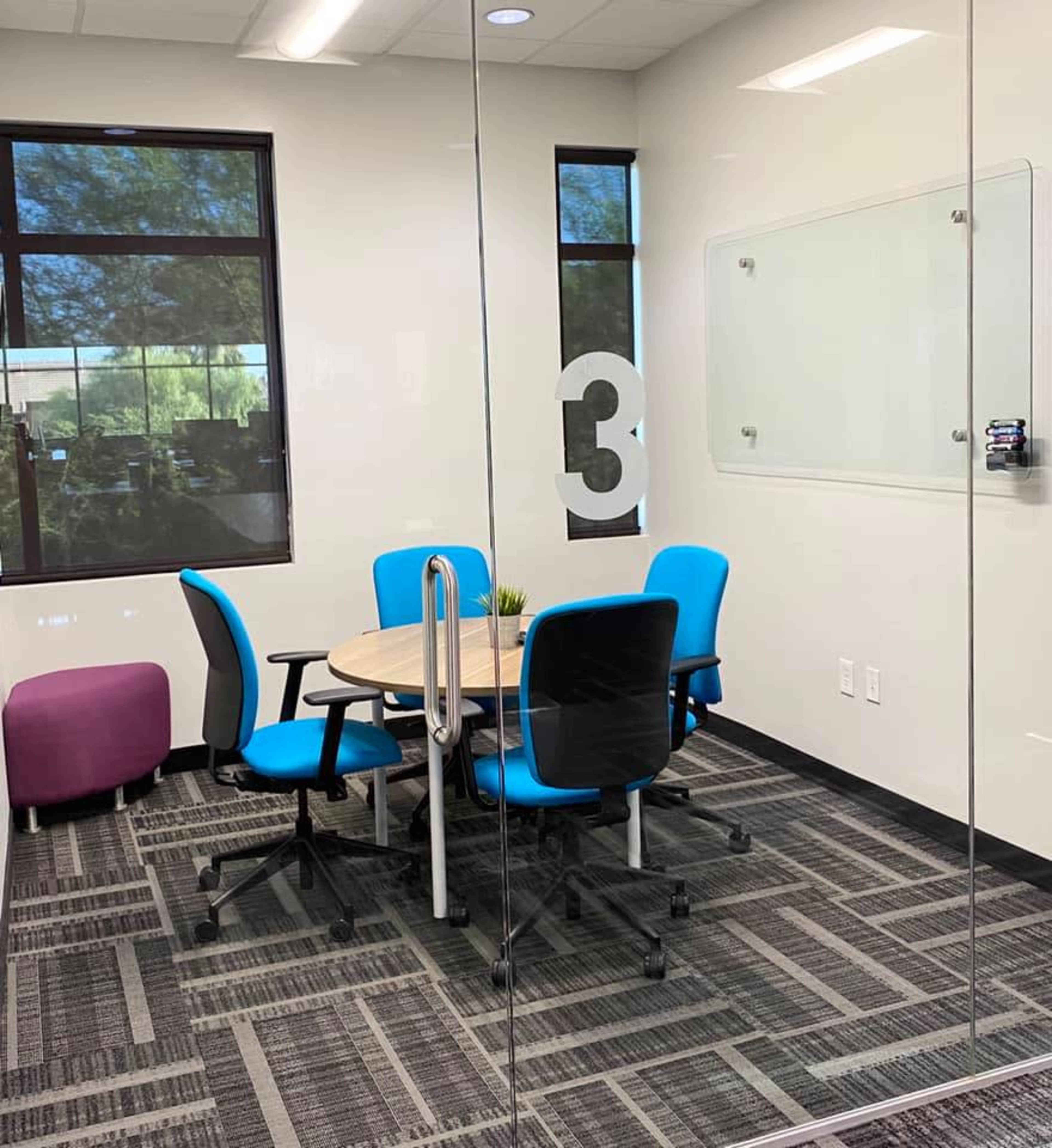 A small conference room features a round table surrounded by four blue chairs and a purple ottoman, with large windows allowing natural light.