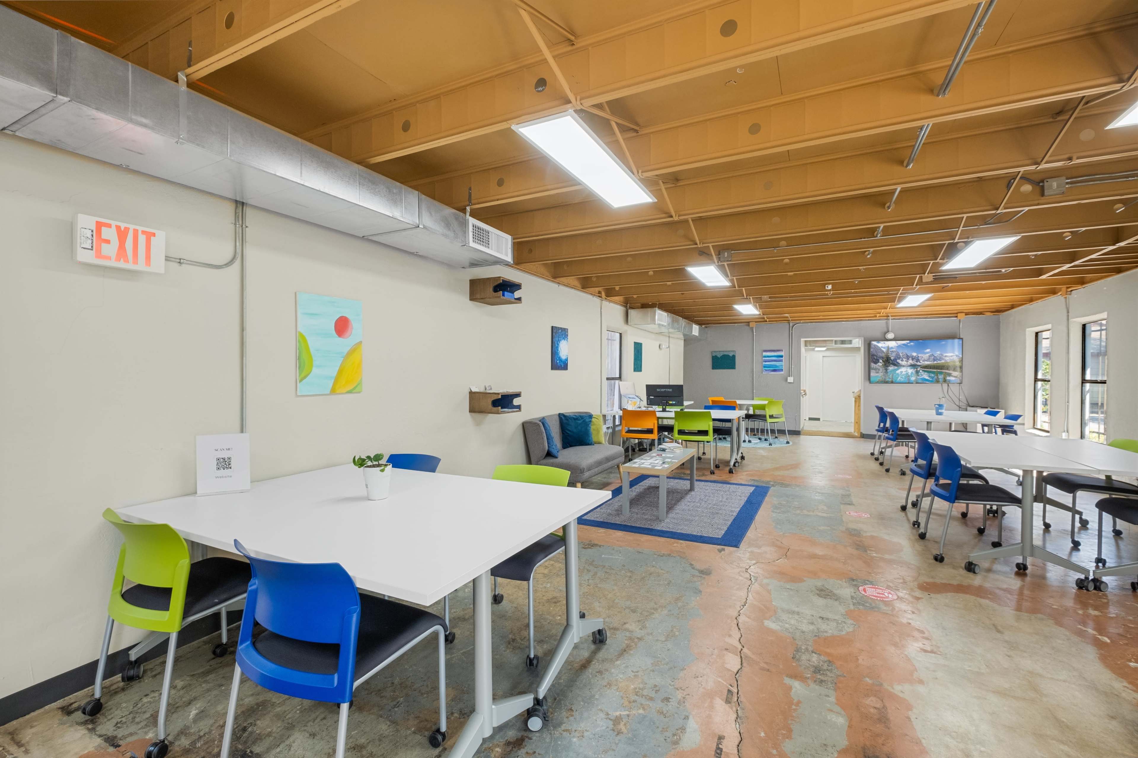 The image shows a spacious, well-lit office area with multiple tables and chairs, a television screen, and colorful wall art.