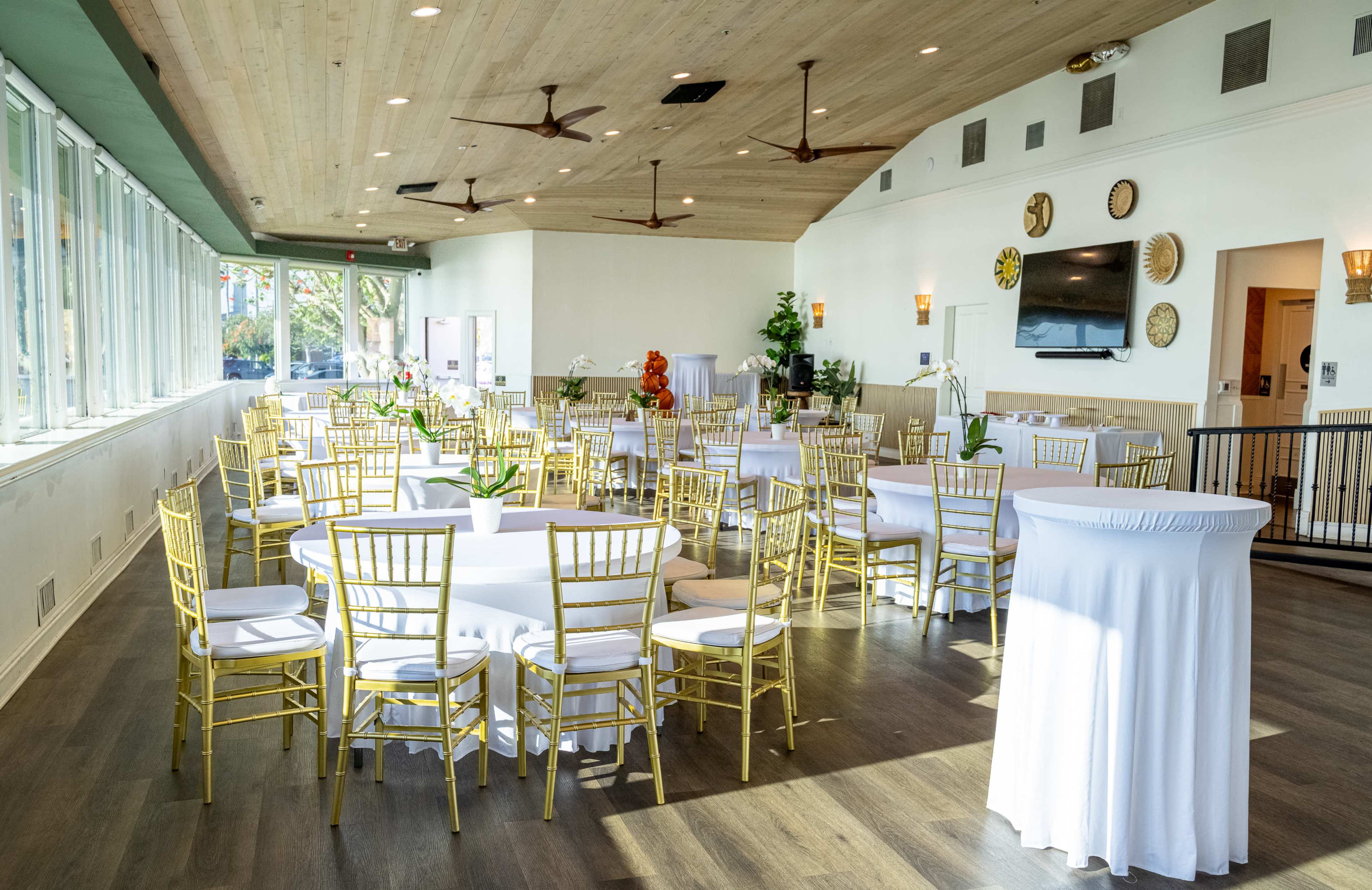 The image shows a spacious event hall set up with gold-framed chairs and white tables, featuring large windows that allow natural light to fill the room.