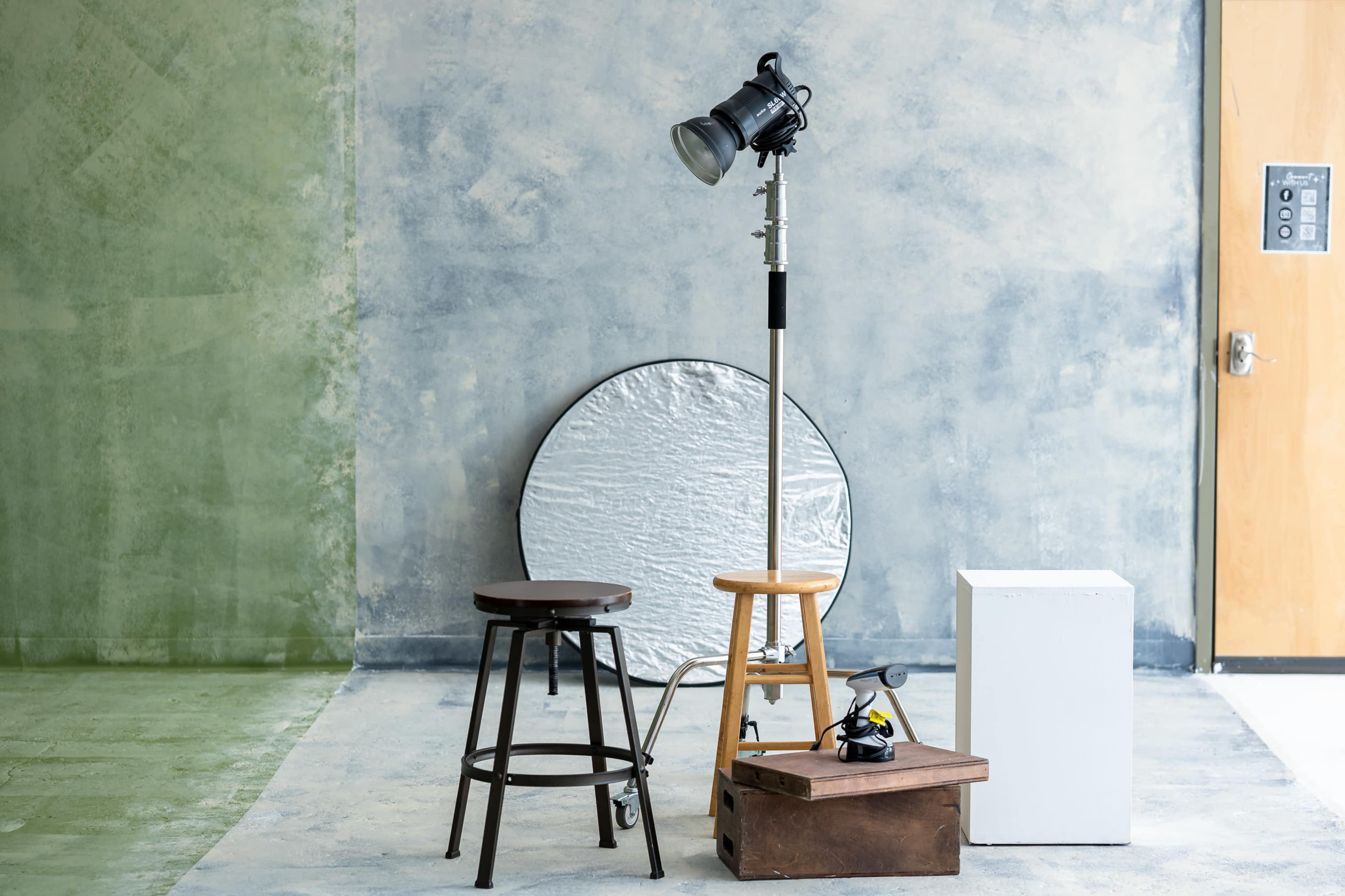 The image features a studio setup with three different stools, a reflective disc, and a spotlight on a stand against a textured wall.