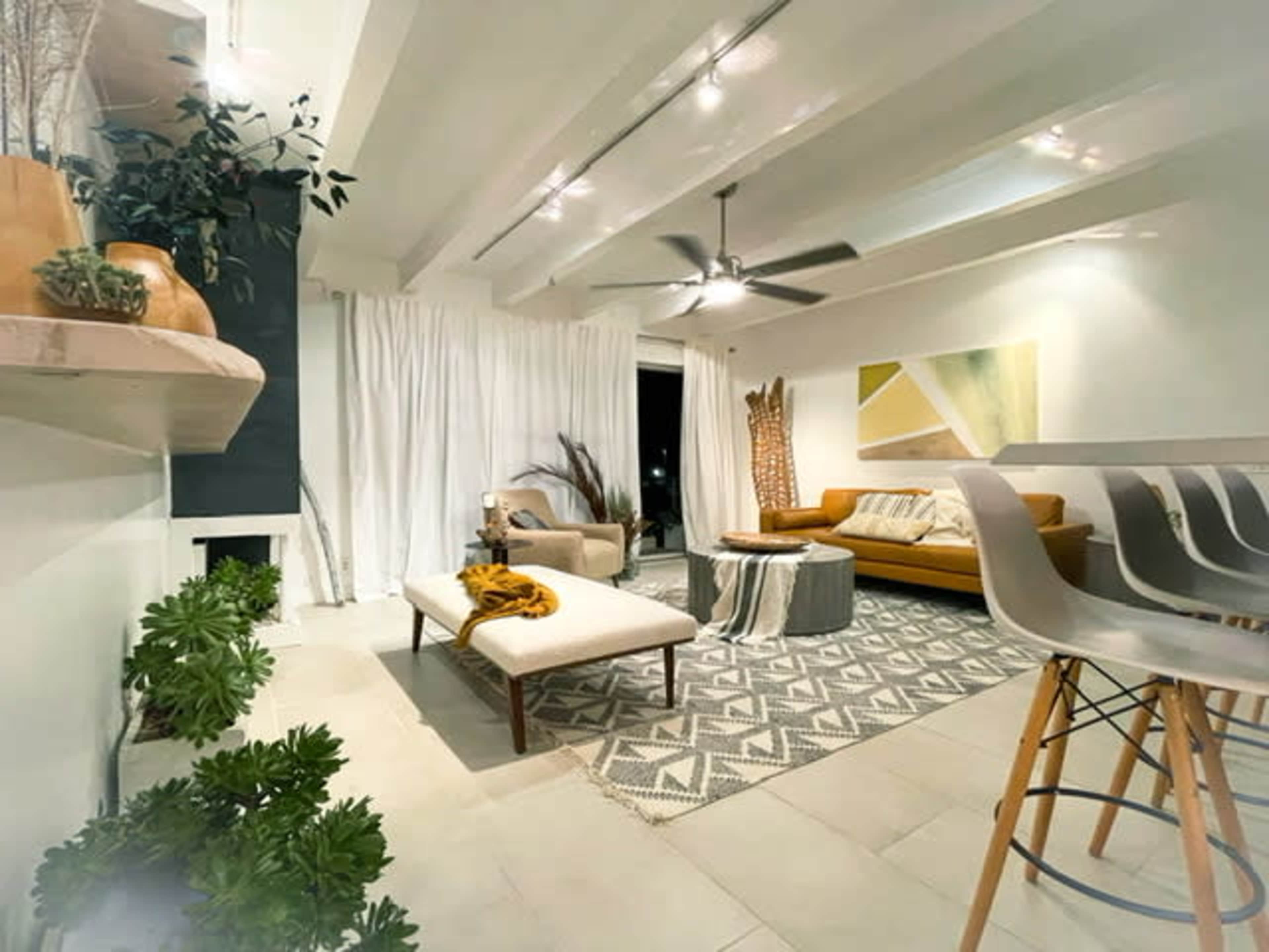 A modern living room with a couch, coffee table, patterned rug, and decorative plants, illuminated by ceiling lights.