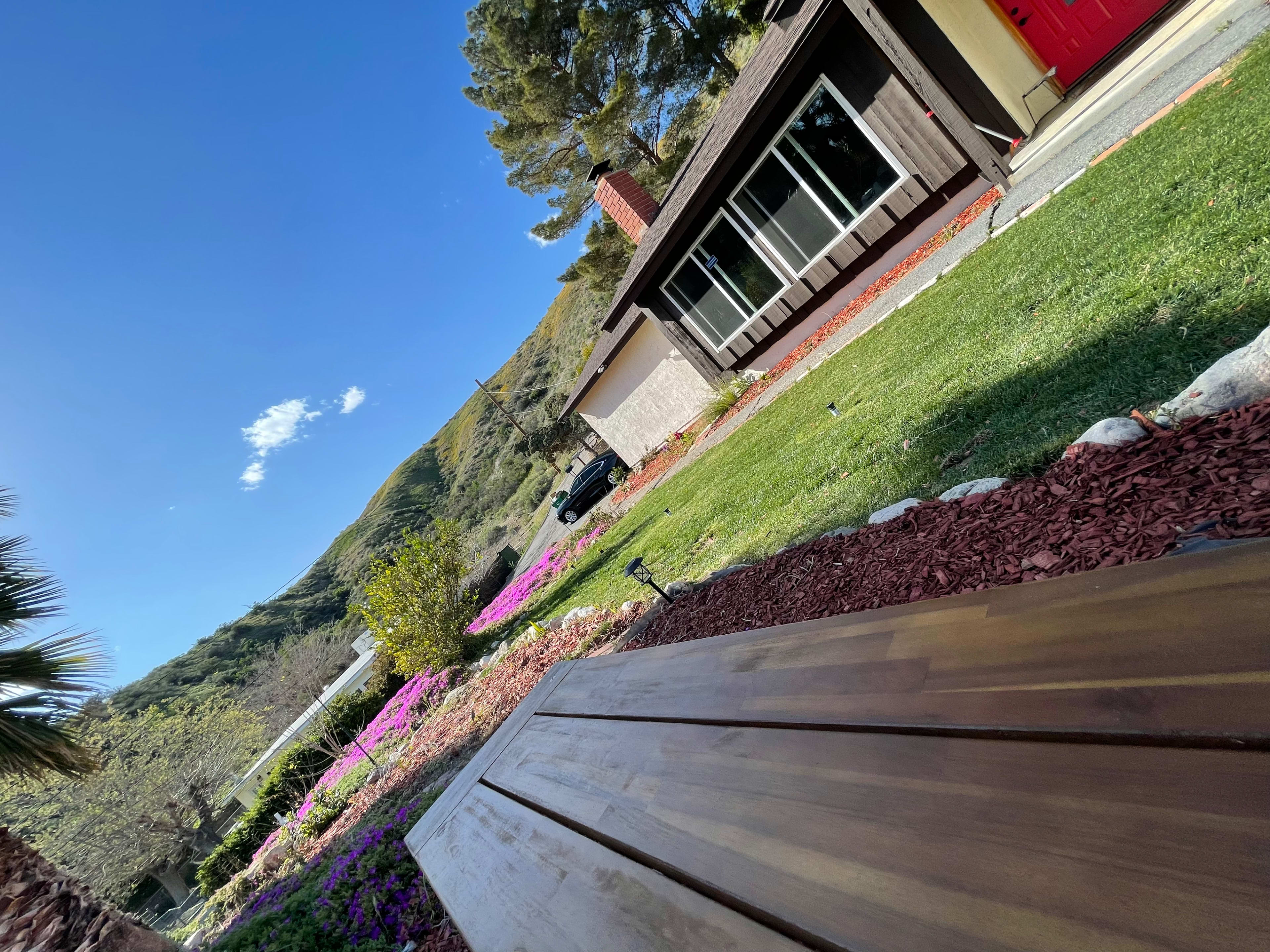 A view of a house surrounded by colorful flowers and a lawn, with mountains in the background under a clear blue sky.