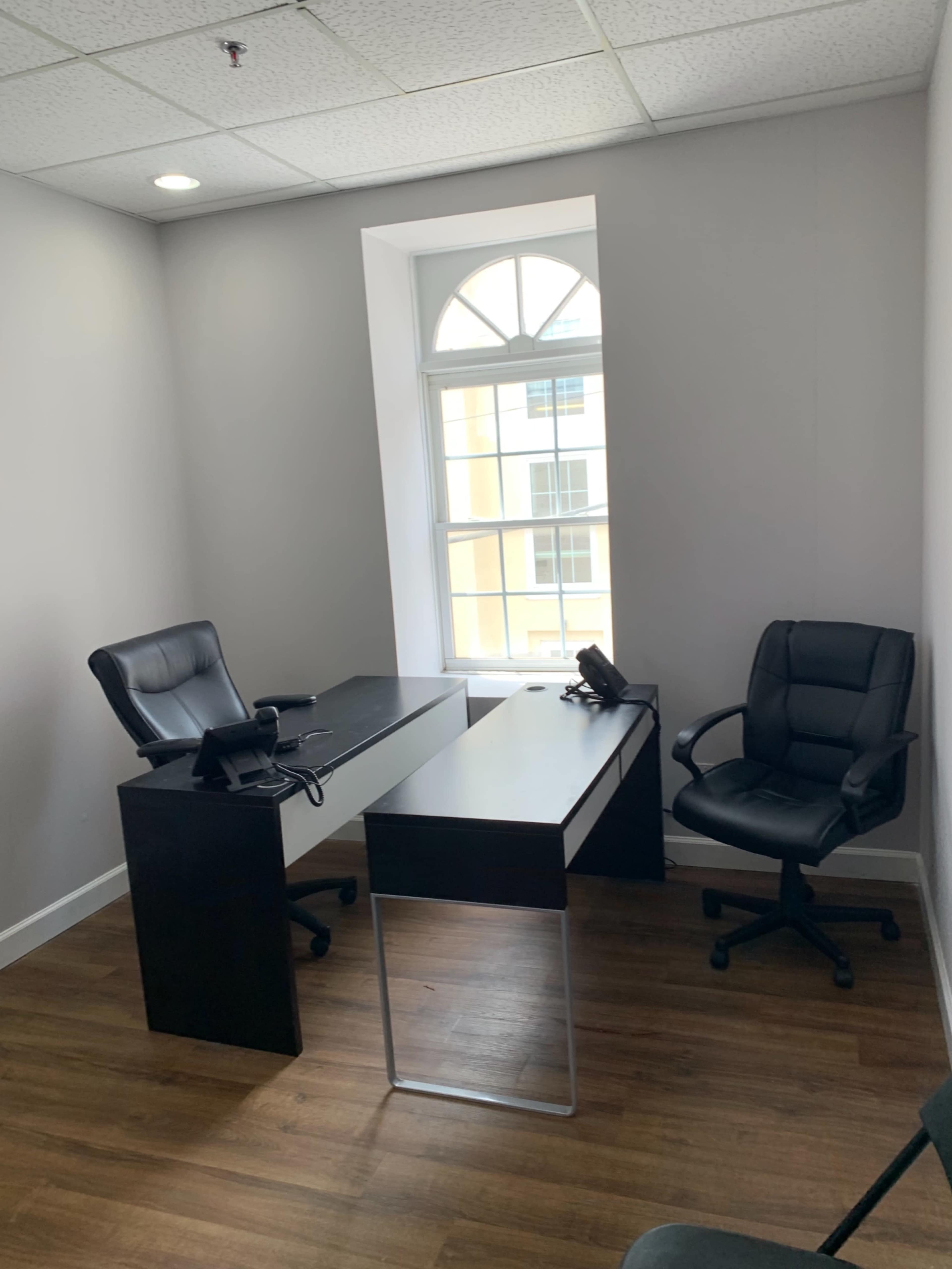 The image shows a small office with a black desk, two black office chairs, and a window letting in natural light.