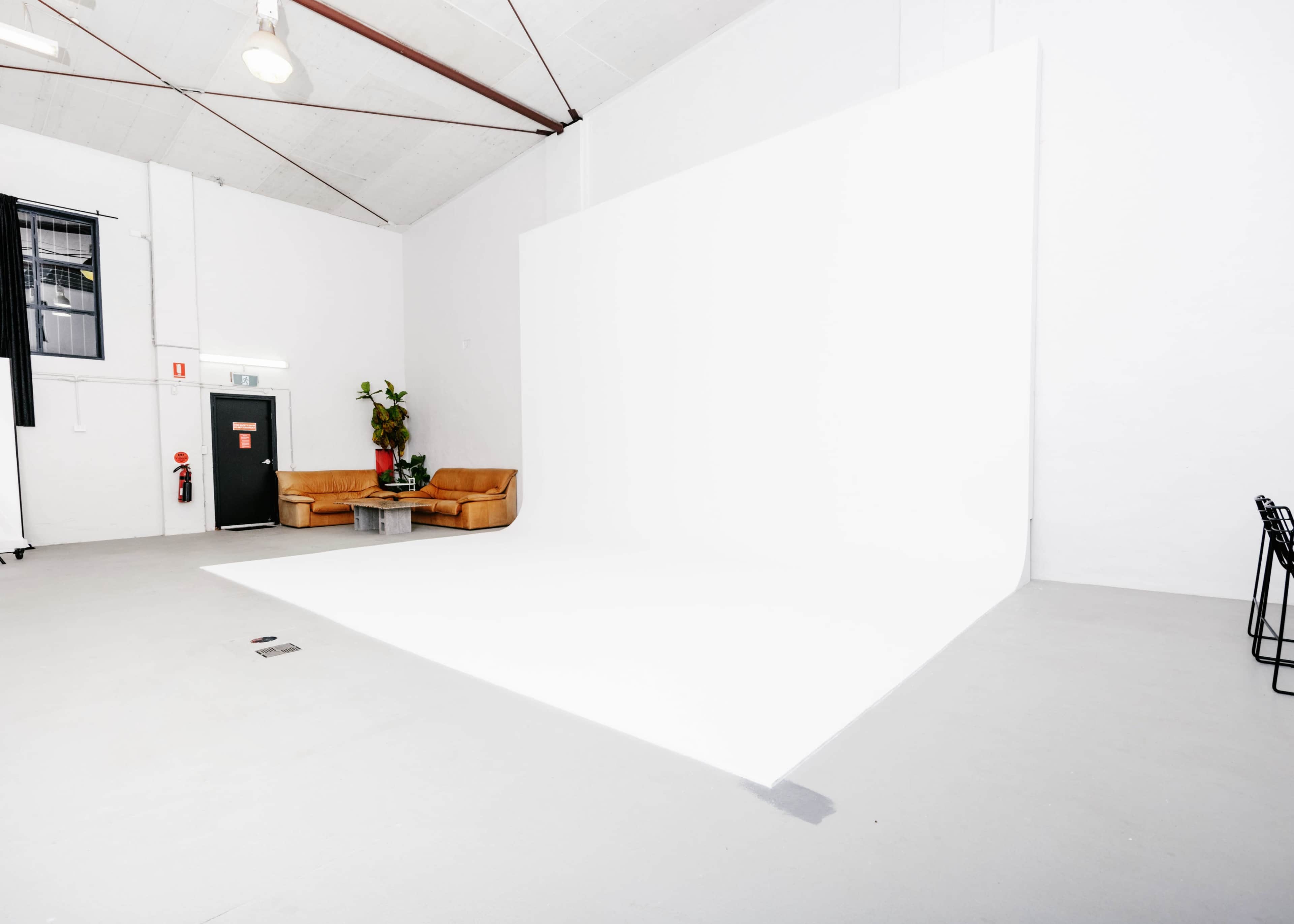 A spacious, minimalist studio with a large white wall and light-colored flooring, featuring a brown couch and a small table.