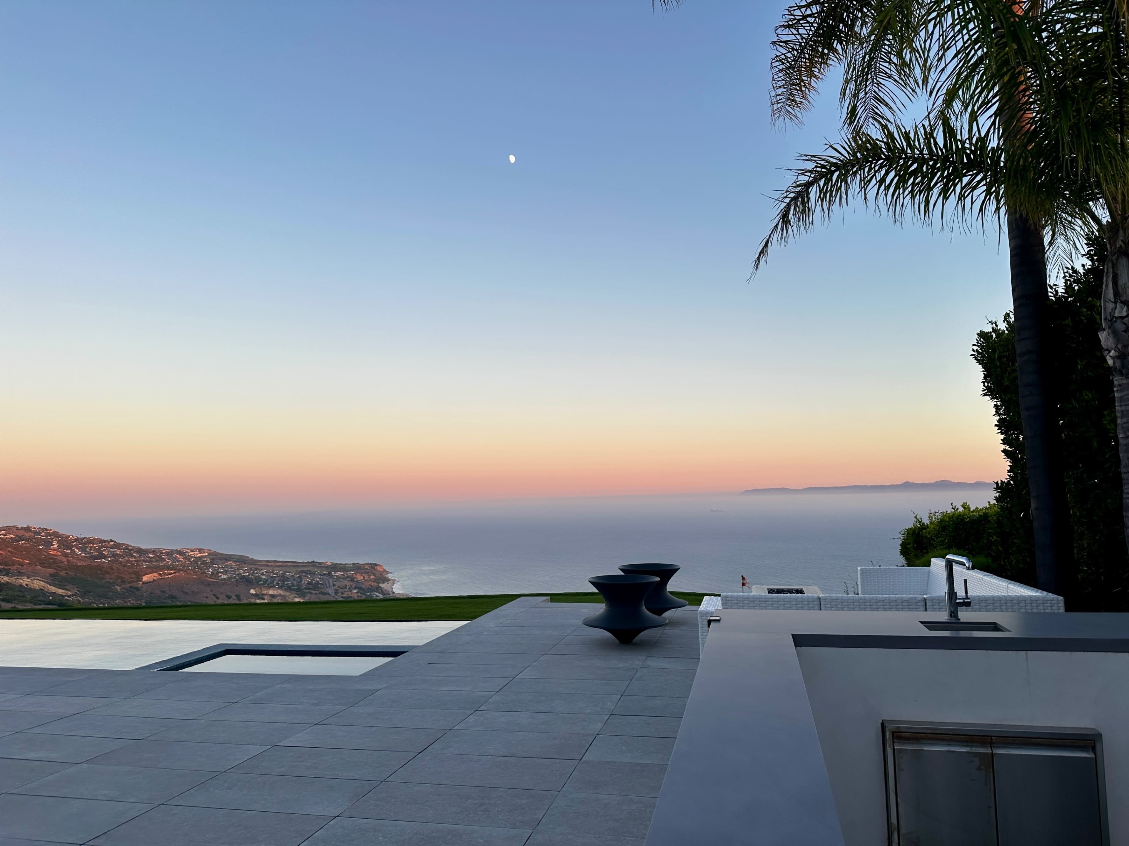 A modern patio with a pool overlooks a coastal landscape at sunset.