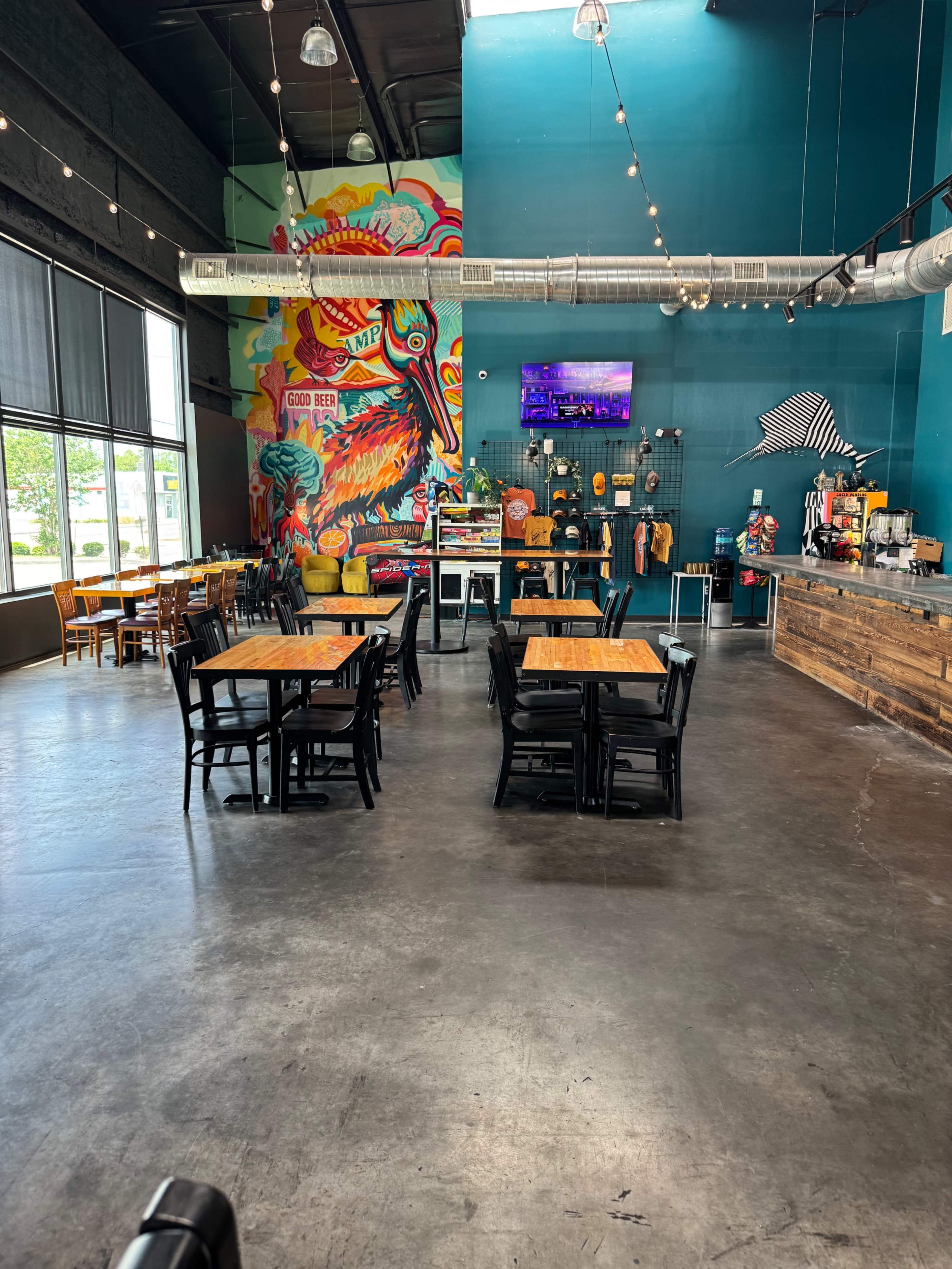 The image shows a brightly colored restaurant interior with wooden tables, black chairs, and a large mural featuring vibrant artwork.