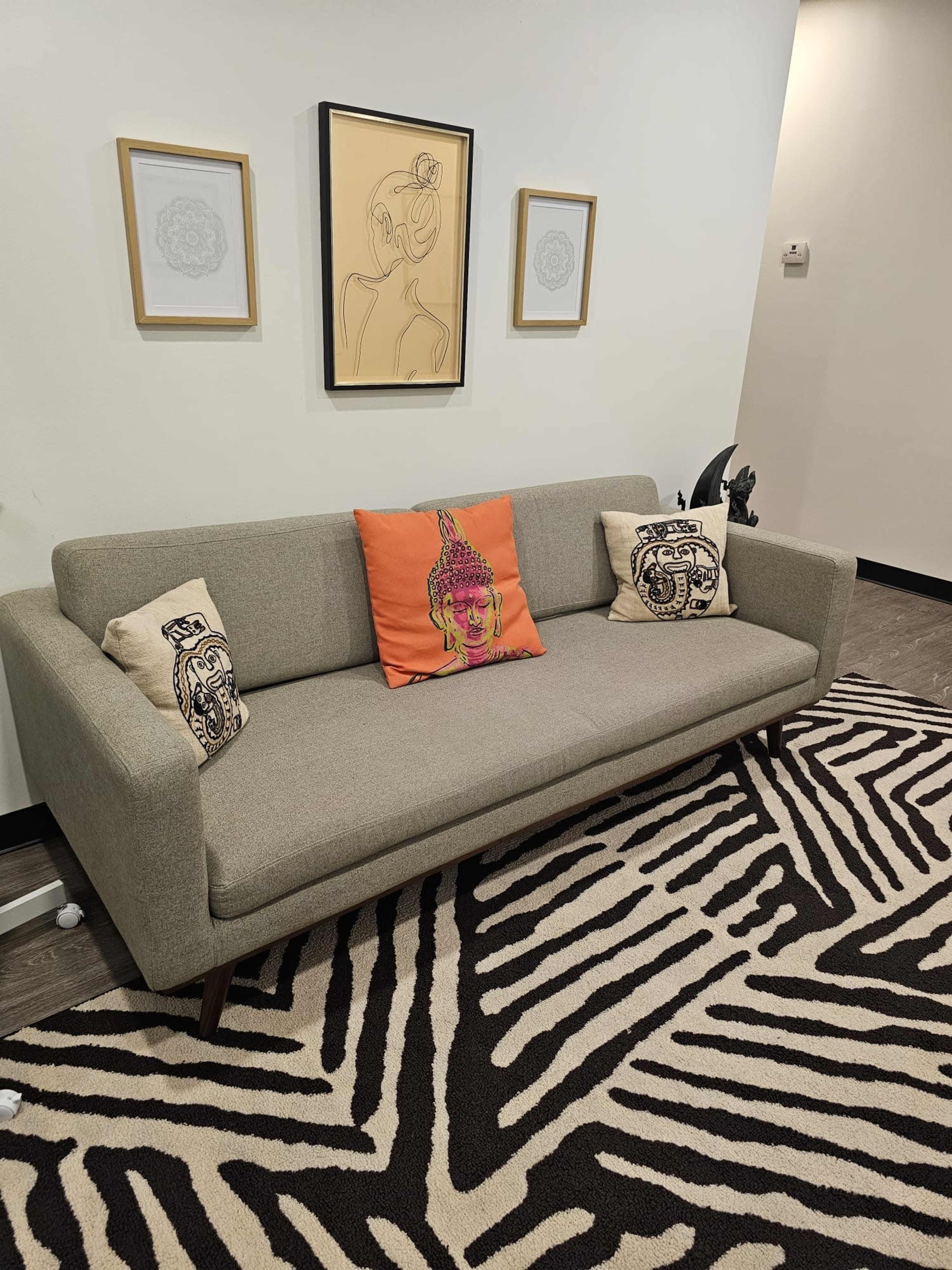 A gray sofa with decorative pillows sits on a zebra-patterned rug in a brightly lit room, with framed artwork hanging above it.