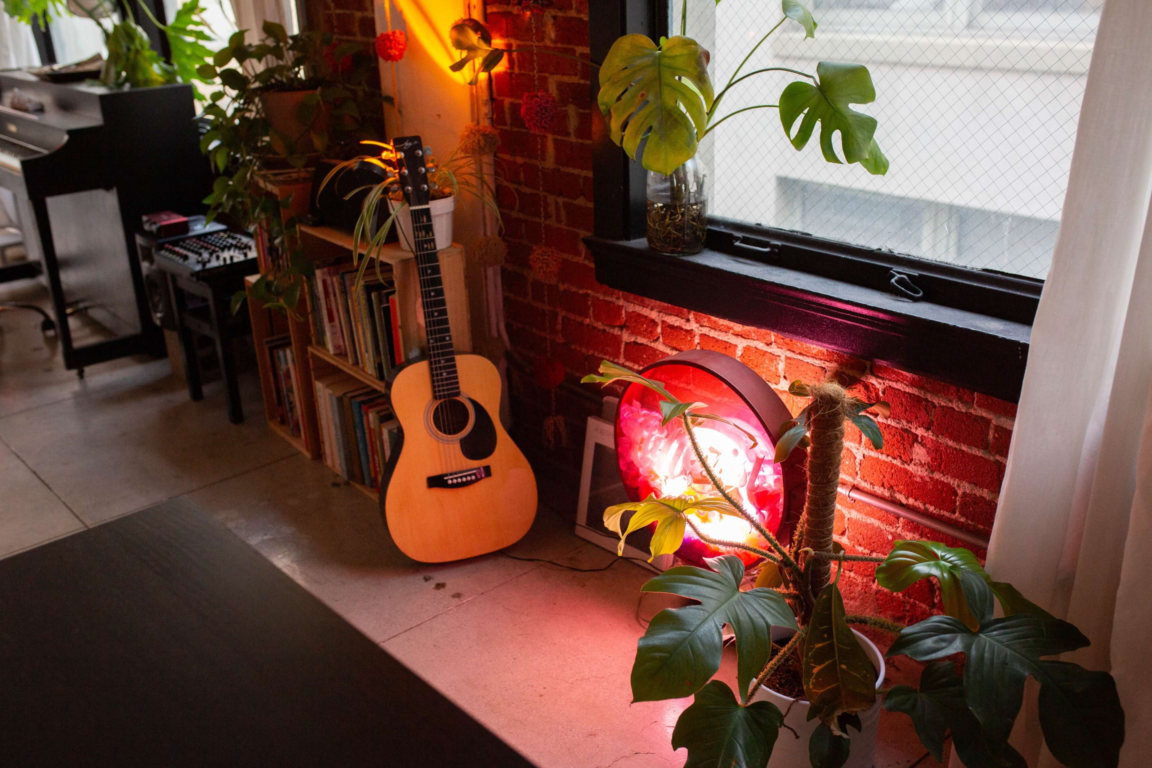 A wooden acoustic guitar leans against a brick wall adorned with various plants and a round light fixture.
