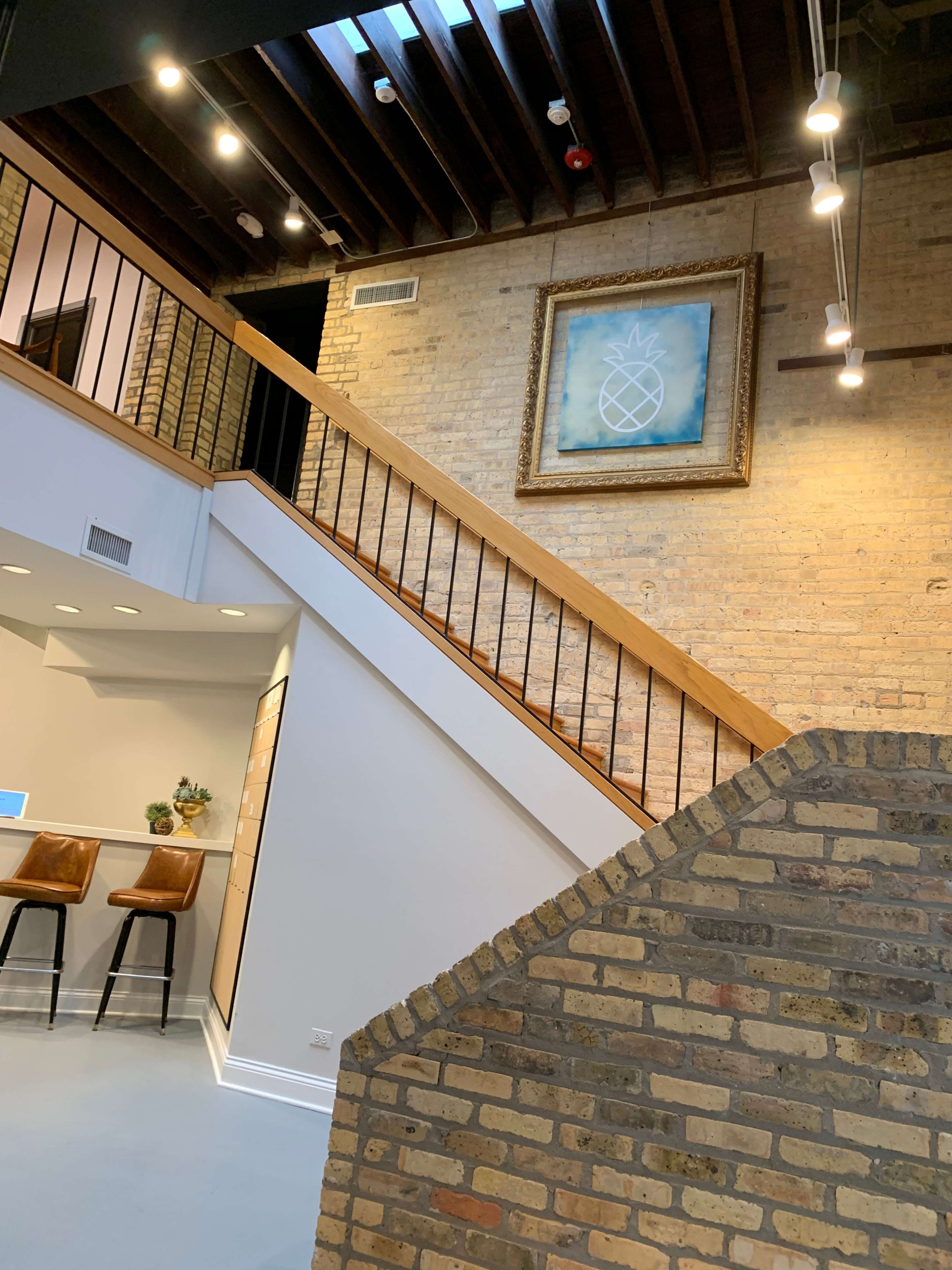 The image shows a modern interior space with a staircase leading to an upper level, featuring a framed pineapple artwork on the wall and bar stools at a counter.